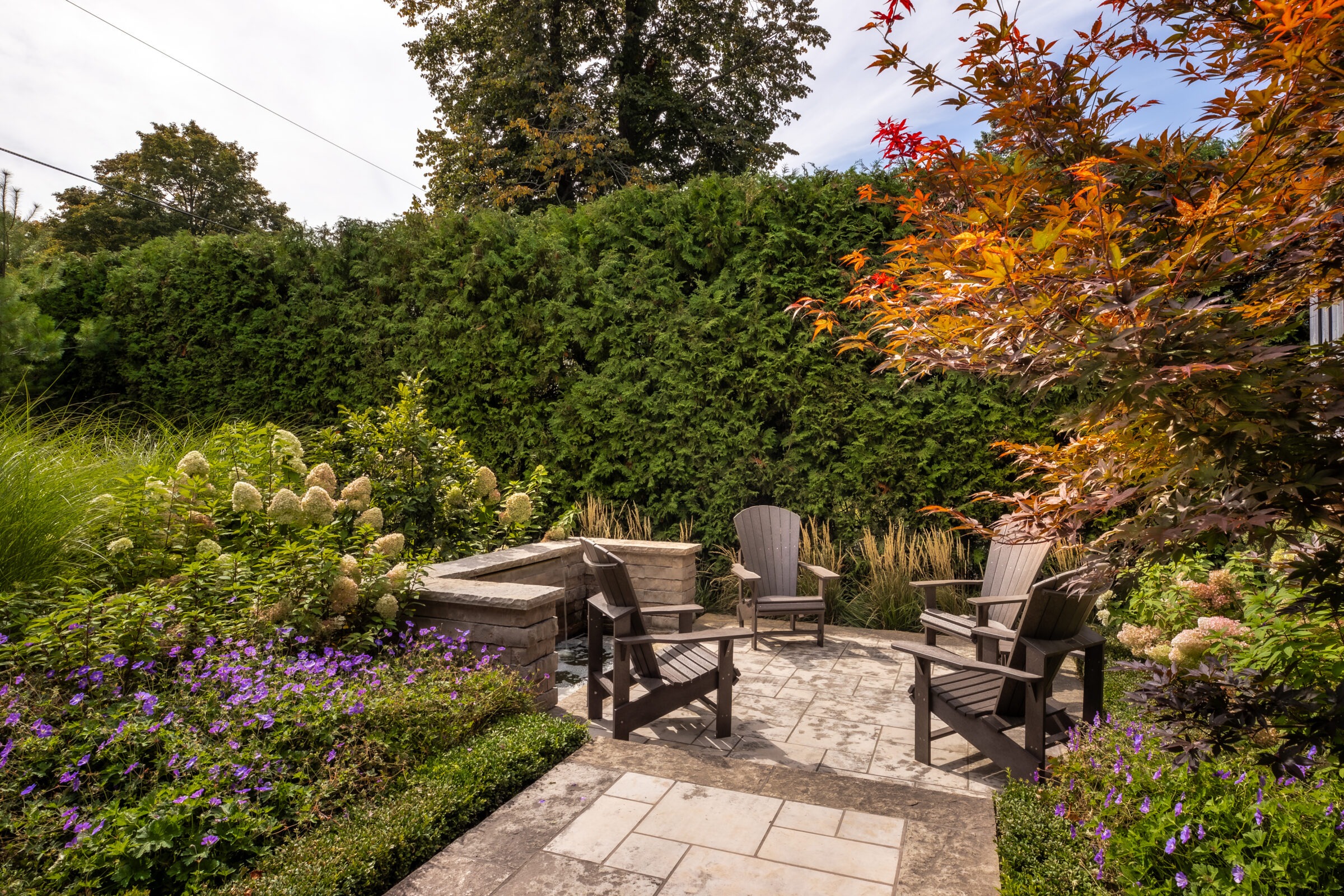 A serene garden patio with wooden chairs, lush greenery, and colorful flowers, surrounded by a tall hedge and shaded by trees.