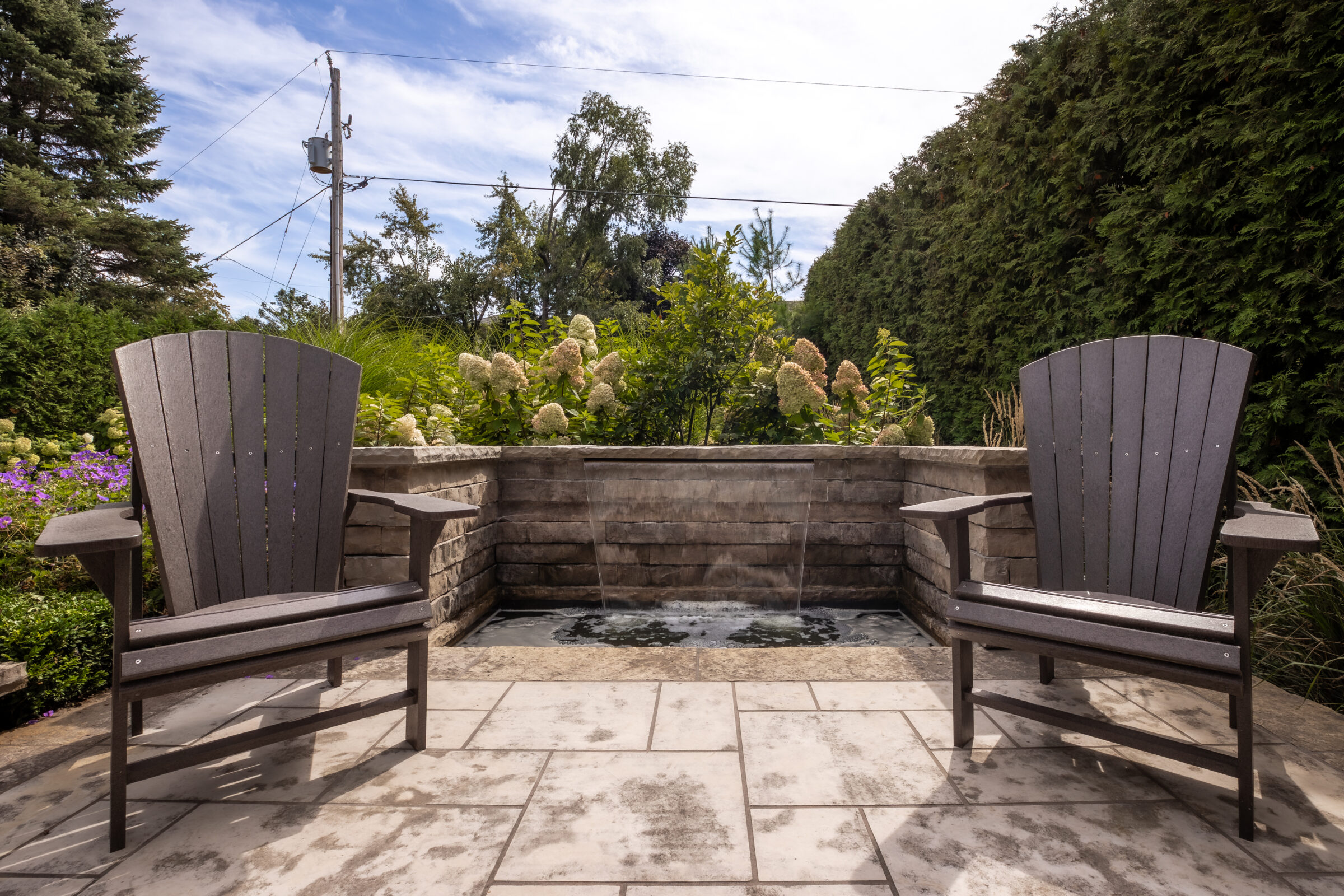 Two wooden chairs on a stone patio face a garden water feature, with lush greenery and blue sky in the background.