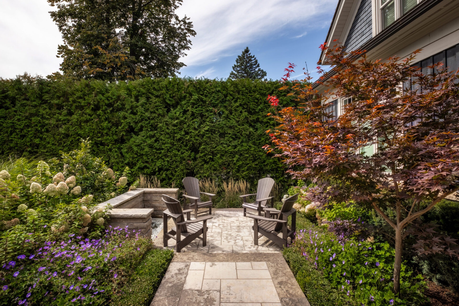 A tranquil garden with Adirondack chairs, vibrant flowers, and a small patio next to a house, surrounded by tall green hedges.