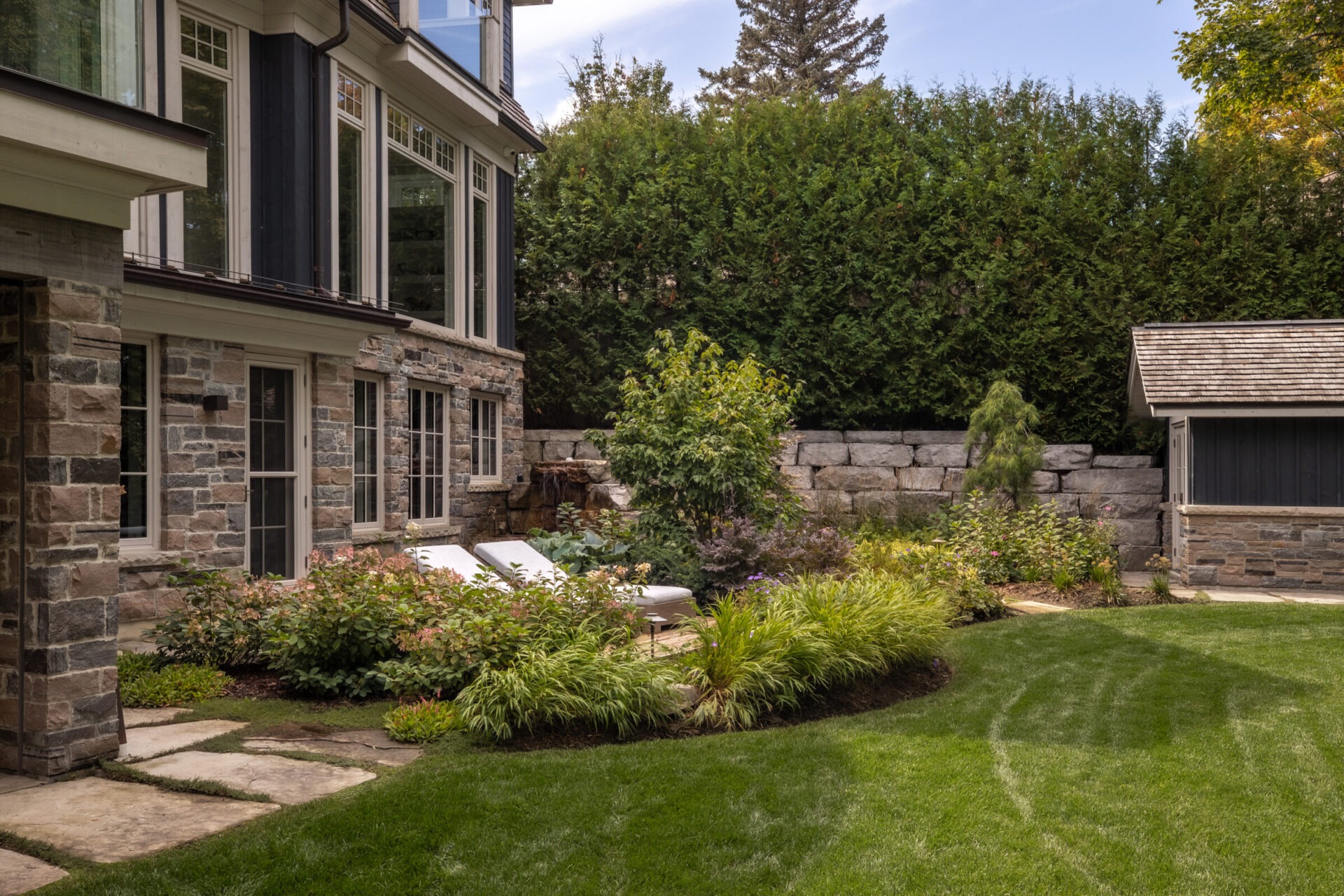 A stone house with large windows overlooks a manicured garden, featuring lush greenery and a small stone outbuilding, surrounded by tall hedges.