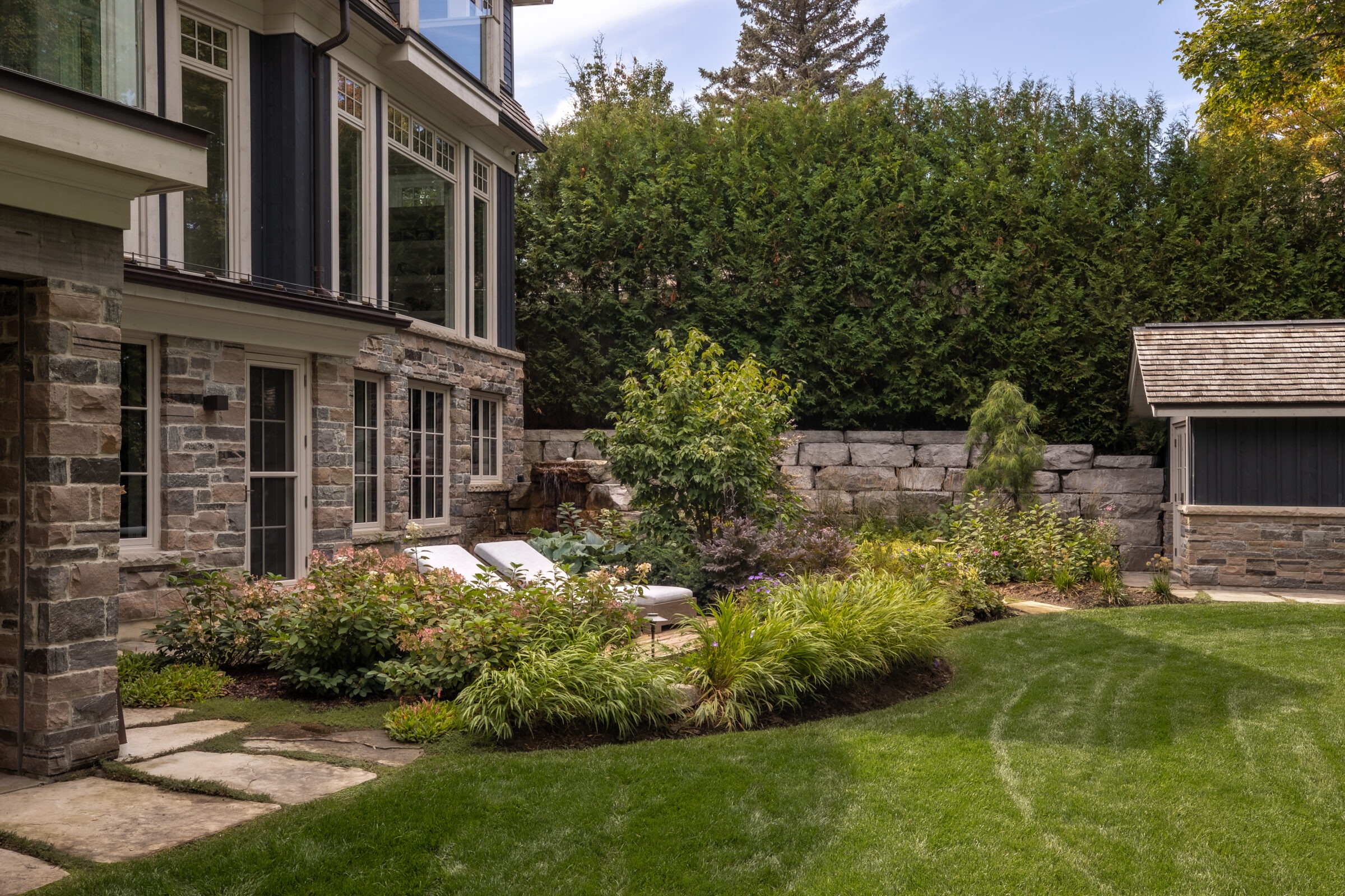 A stone house with large windows overlooks a manicured garden, featuring lush greenery and a small stone outbuilding, surrounded by tall hedges.