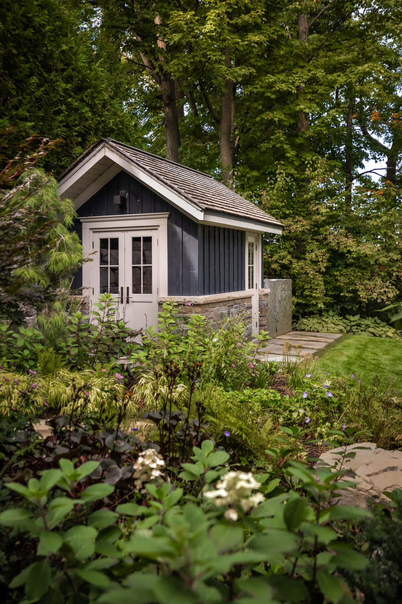 A quaint blue garden shed with white trim is surrounded by lush green foliage and blooming plants, set amidst tall trees.