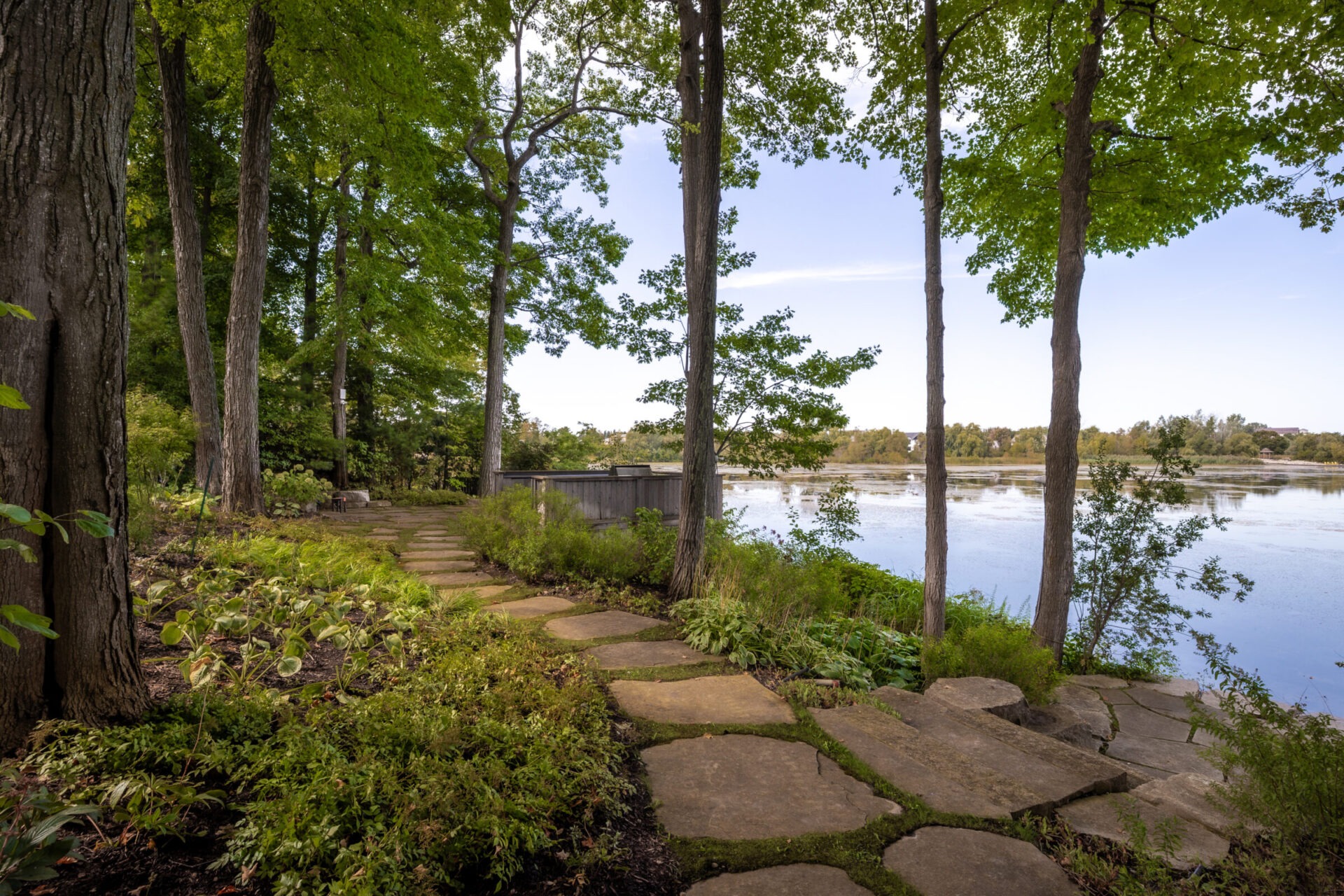 A scenic lakeside view with a stone path winds through tall trees, offering a serene, natural setting by the calm water.