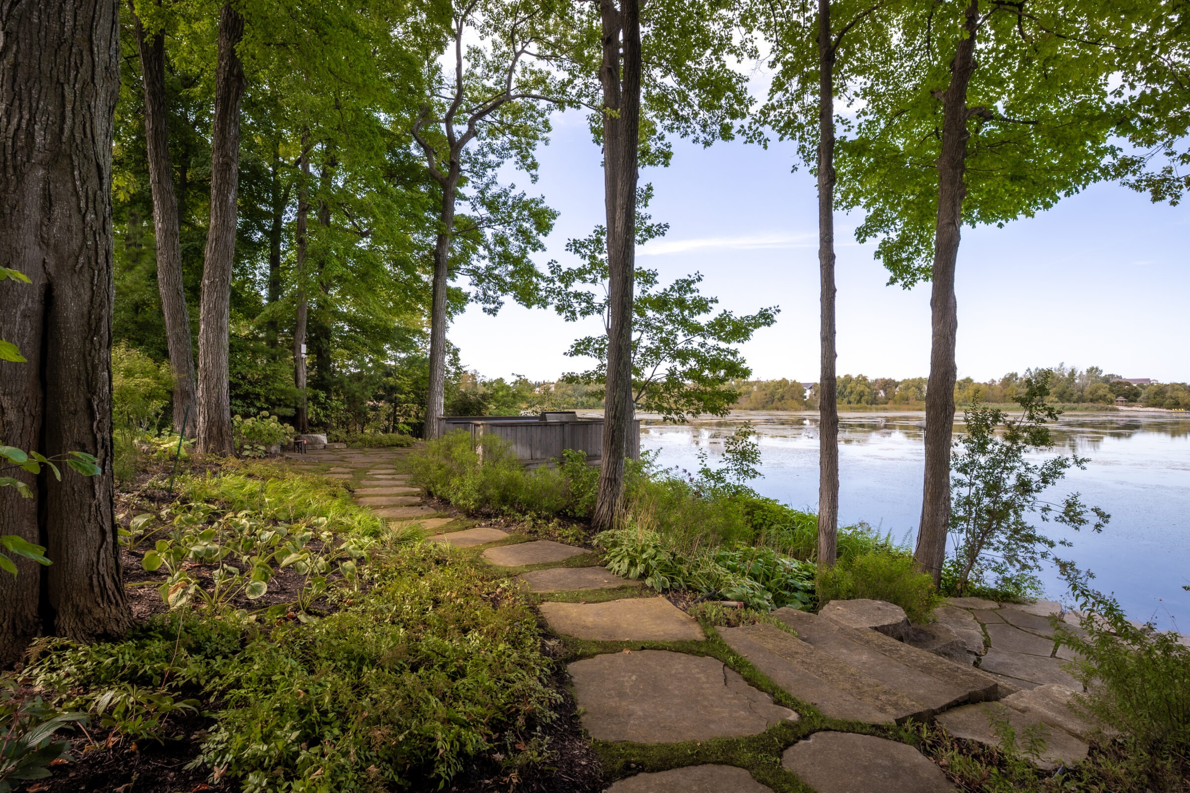 A scenic lakeside view with a stone path winds through tall trees, offering a serene, natural setting by the calm water.