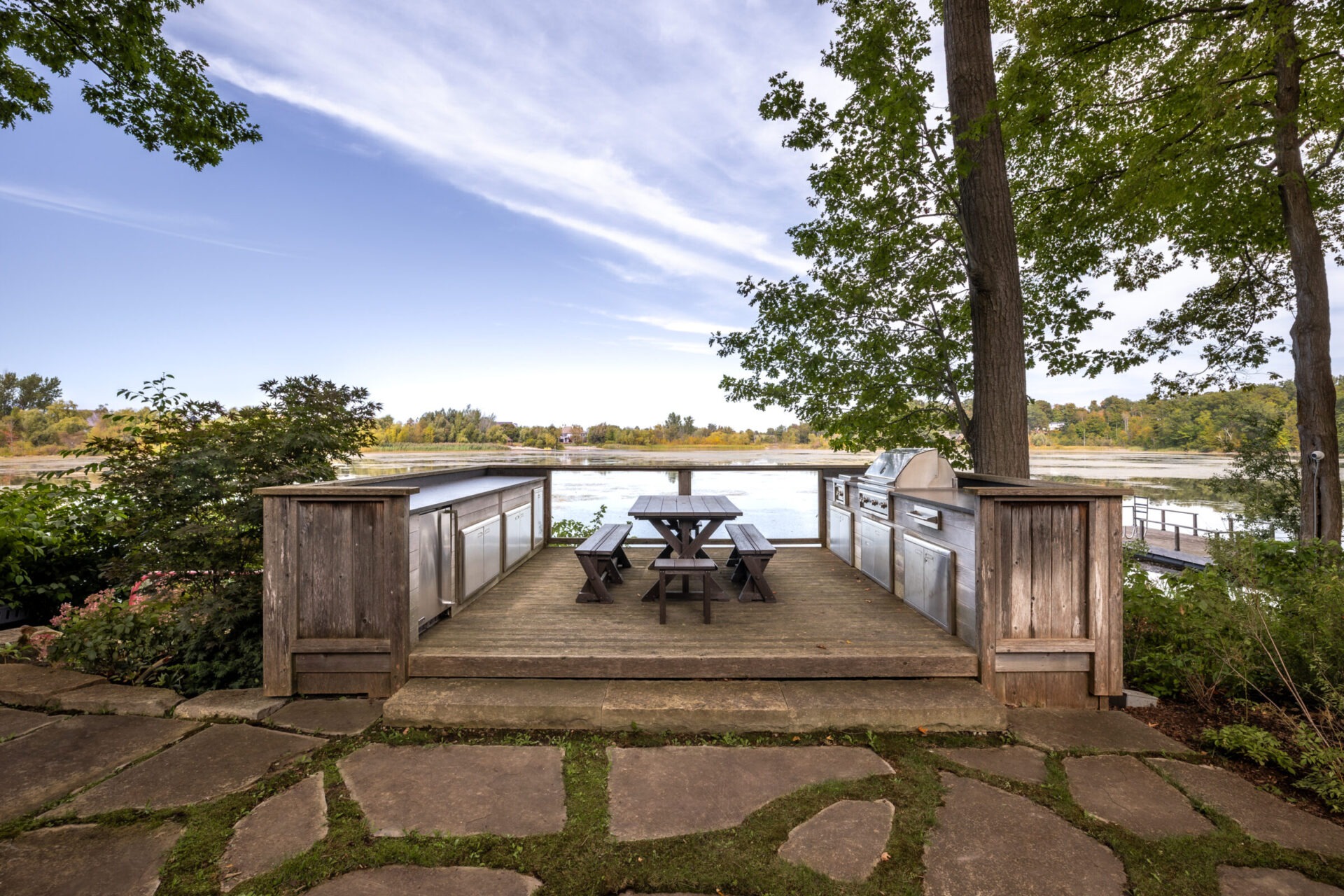 Wooden deck with picnic table and benches overlooking a serene lake, surrounded by trees and stone path, under a clear blue sky.