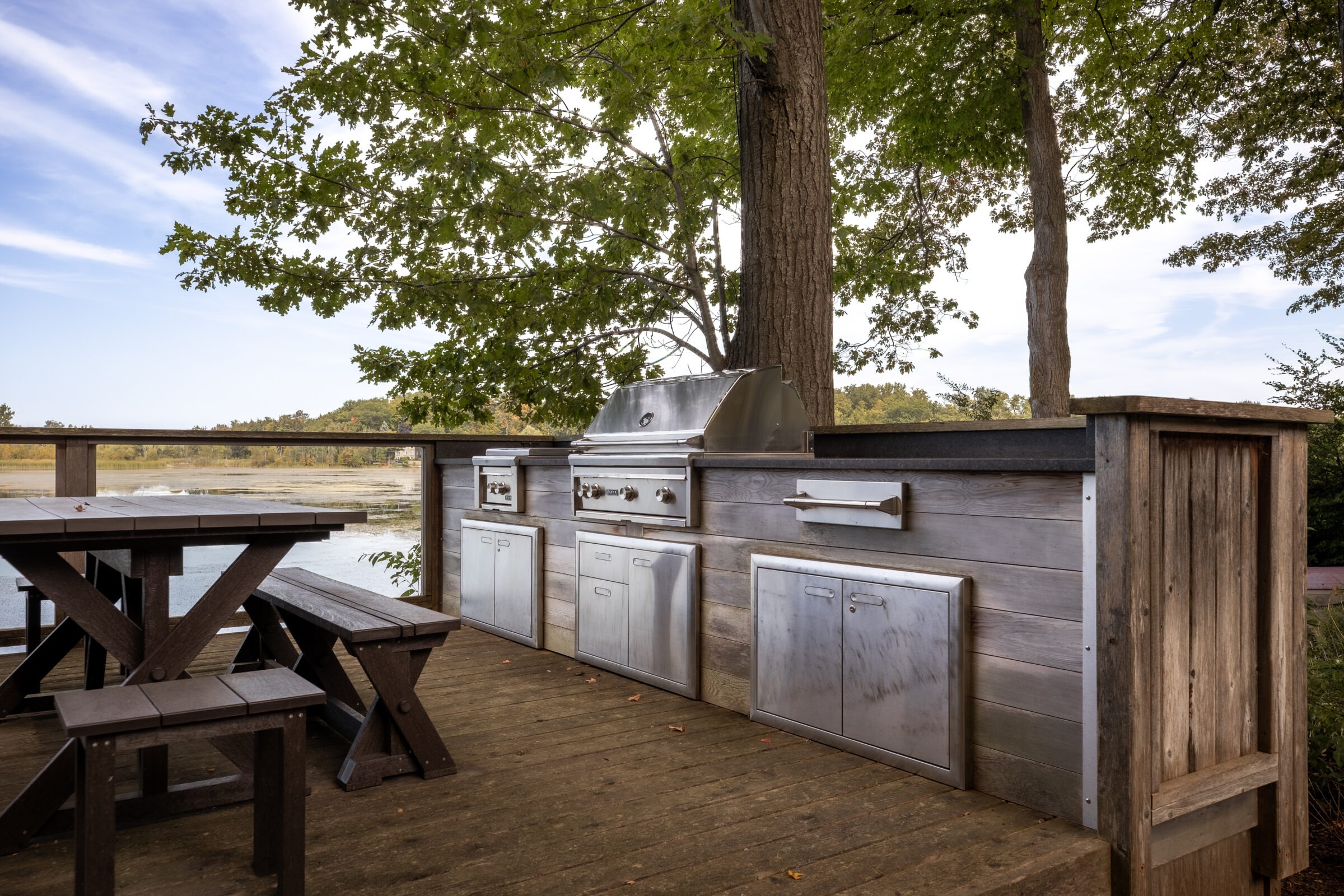Outdoor wooden deck with built-in stainless steel grill, surrounded by trees. Picnic table present. Scenic view of water and greenery. Peaceful setting.