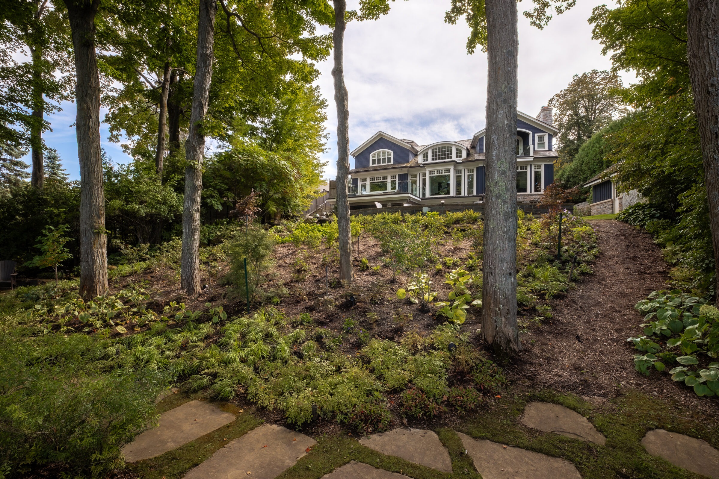 A house with large windows stands on a hill surrounded by trees and a landscaped garden, under a partly cloudy sky.