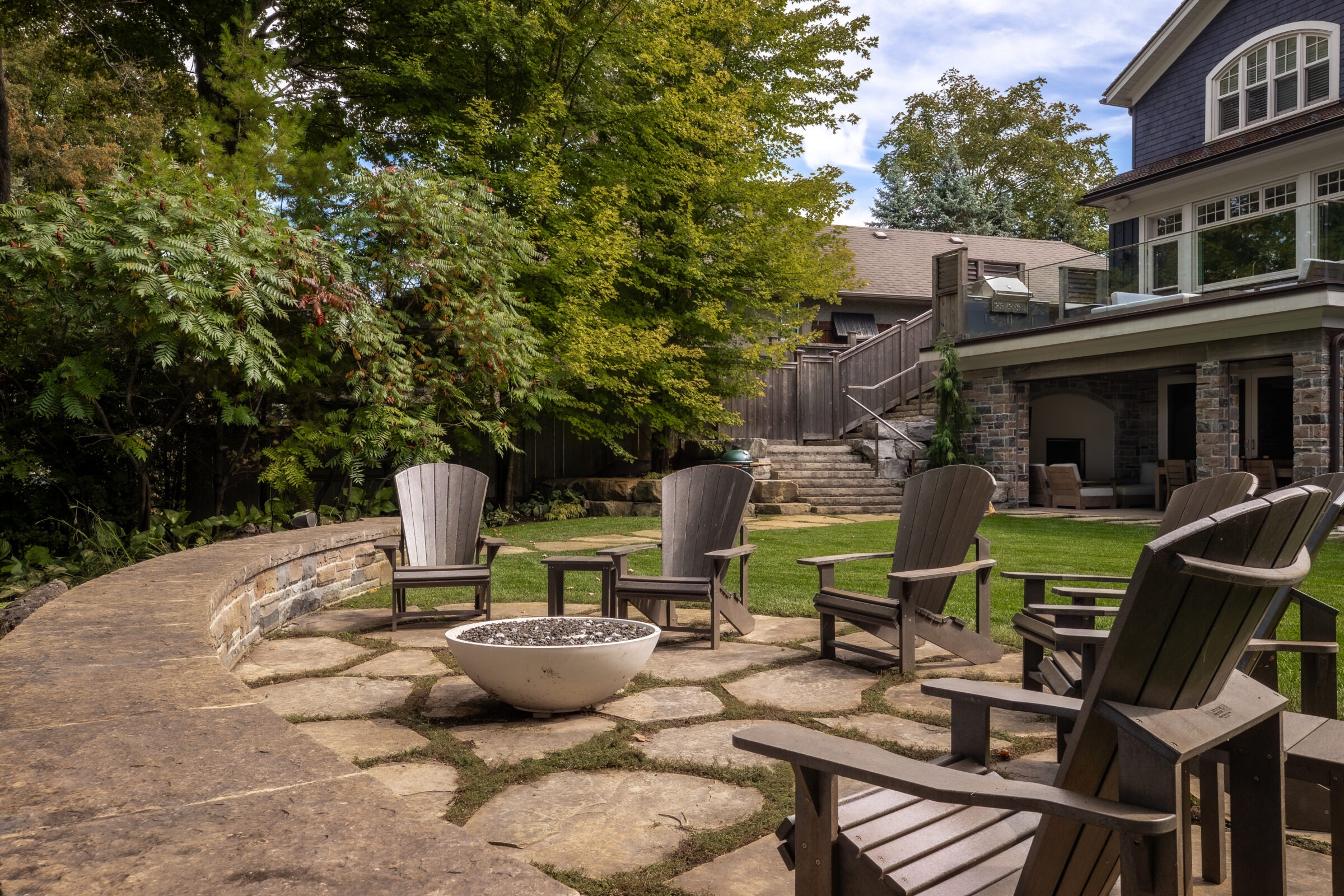 A backyard patio with wooden chairs surrounds a fire pit, near a large stone house and lush greenery under a clear sky.