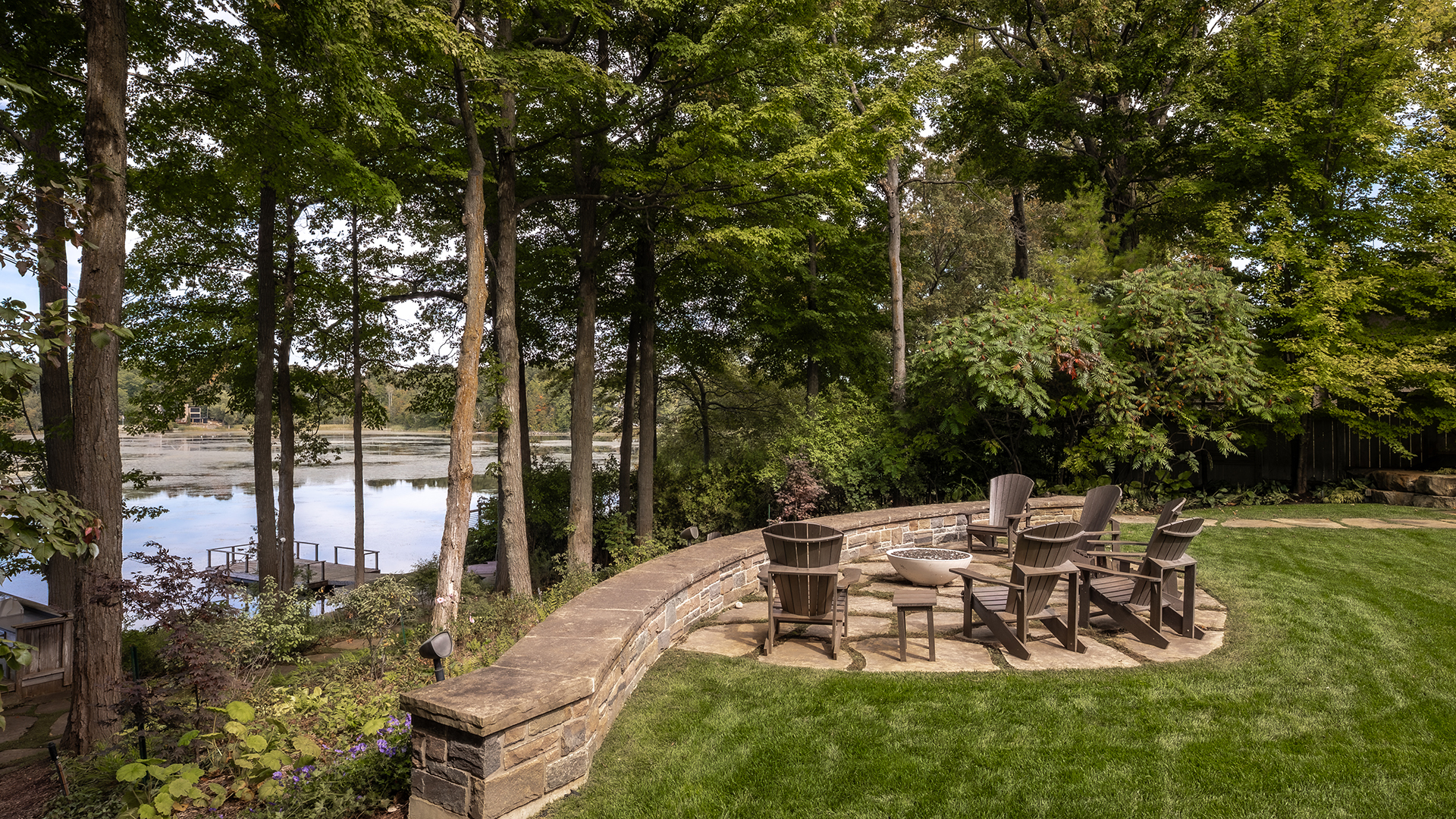 Chairs arranged around a stone patio, beside a lush lawn with tall trees and a serene lake view in the background.