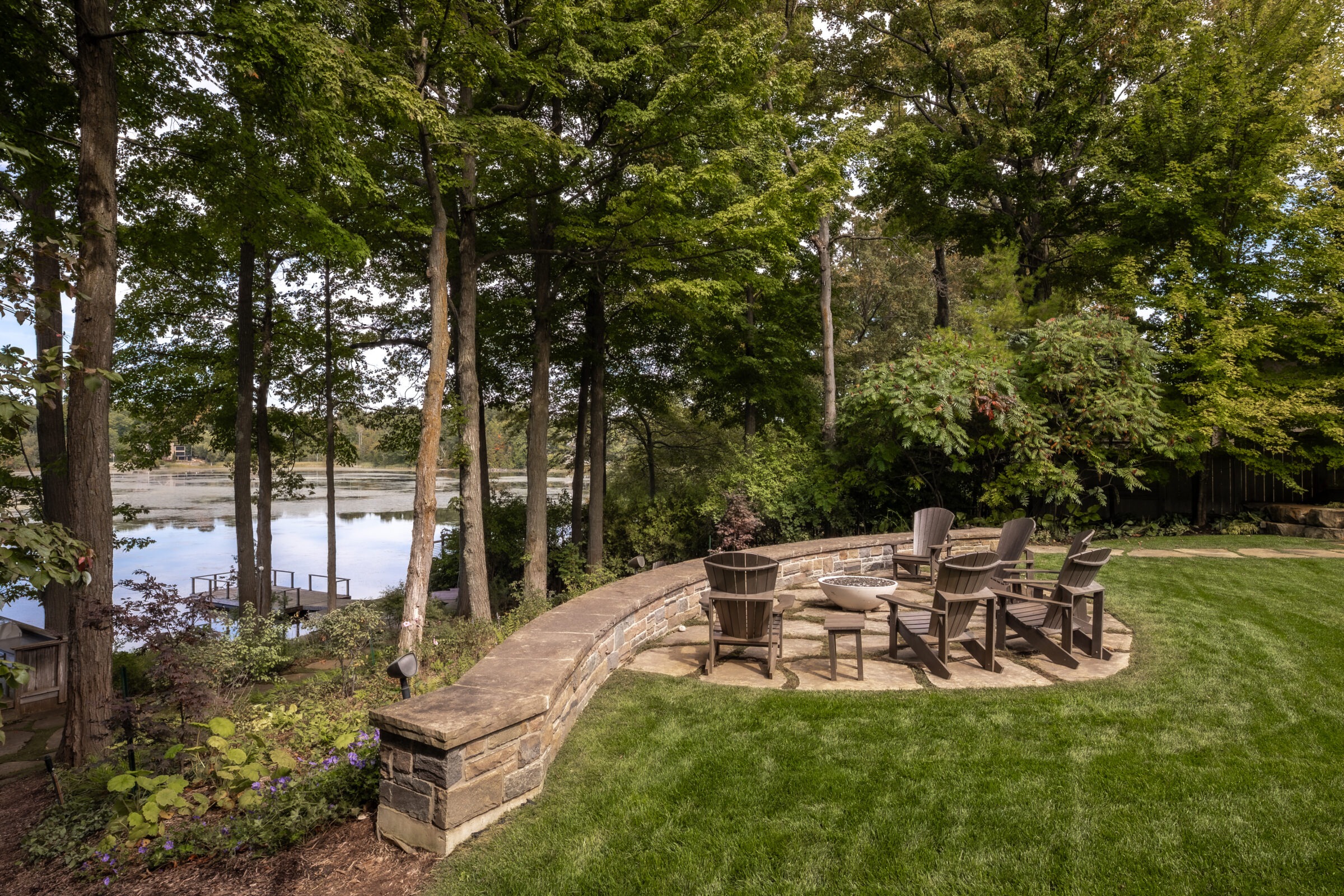 A serene backyard features a stone patio with chairs, surrounded by lush trees and overlooking a calm, reflective lake.