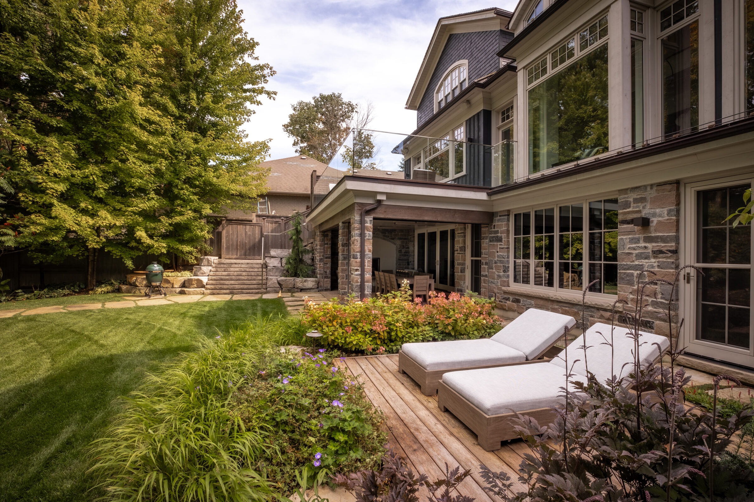 Elegant backyard with lush landscaping, wooden deck, and two lounge chairs. Large house with stone façade and expansive windows in the background.