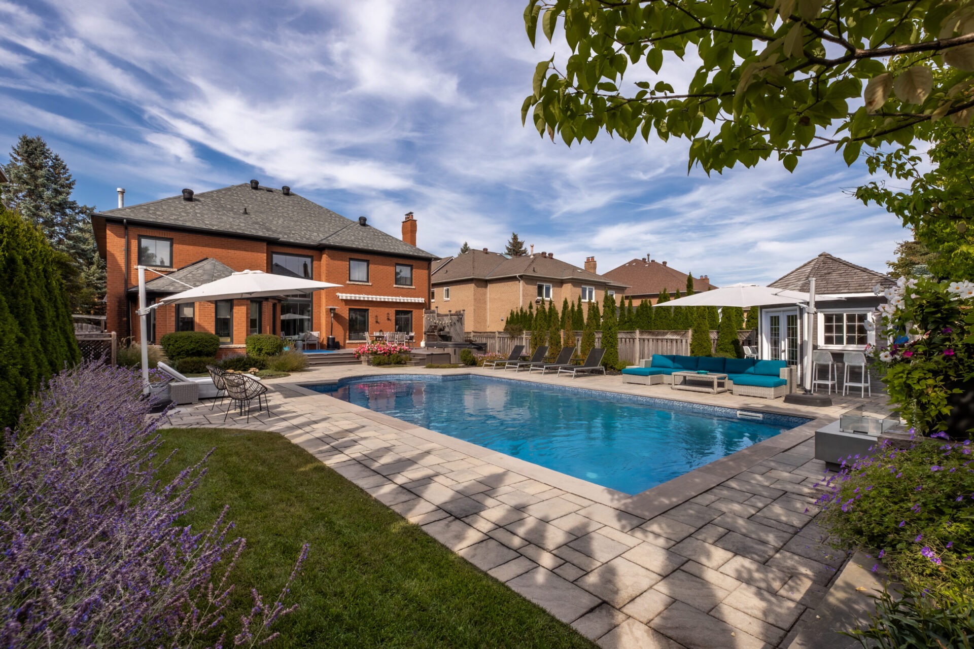 A backyard features a swimming pool surrounded by patio furniture, greenery, and a gazebo, adjacent to a large brick house under a blue sky.