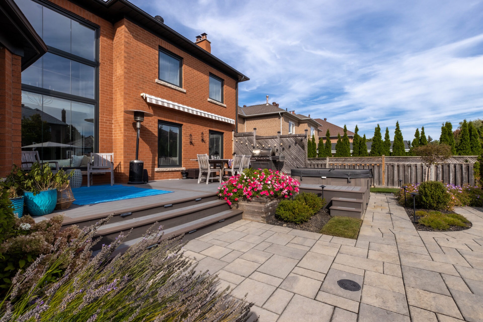 Modern brick house with large windows, a wooden deck, patio furniture, vibrant flowers, and a neatly paved backyard, under a partly cloudy sky.