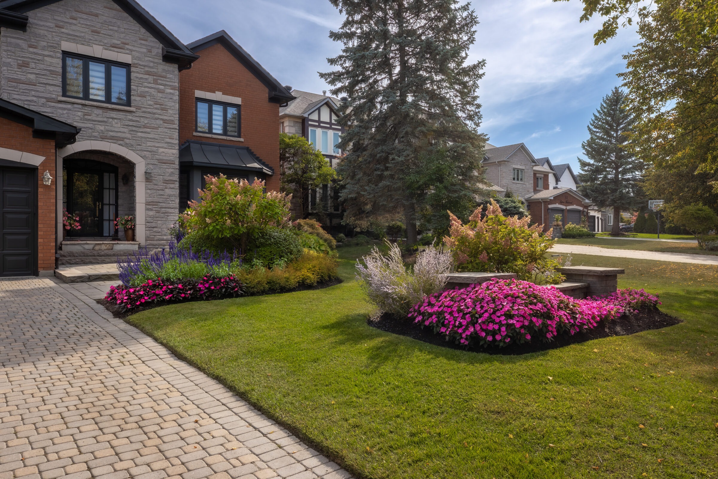 Charming suburban neighborhood with well-maintained lawns and vibrant flowerbeds. Elegant houses line the street under a partly cloudy sky and tall trees.
