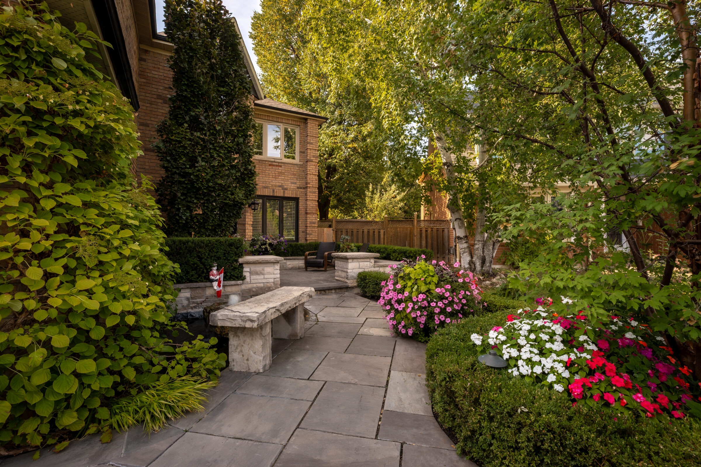A serene garden with stone benches, colorful flowers, and lush greenery beside a red-brick house. Wooden fence and mature trees in the background.