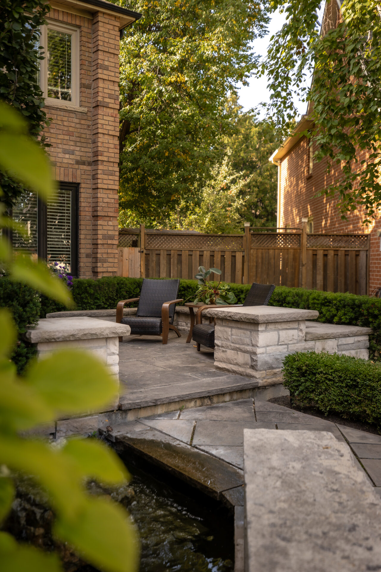 A cozy, private backyard patio with wicker chairs, stone planters, lush greenery, and a tranquil water feature, surrounded by brick houses.