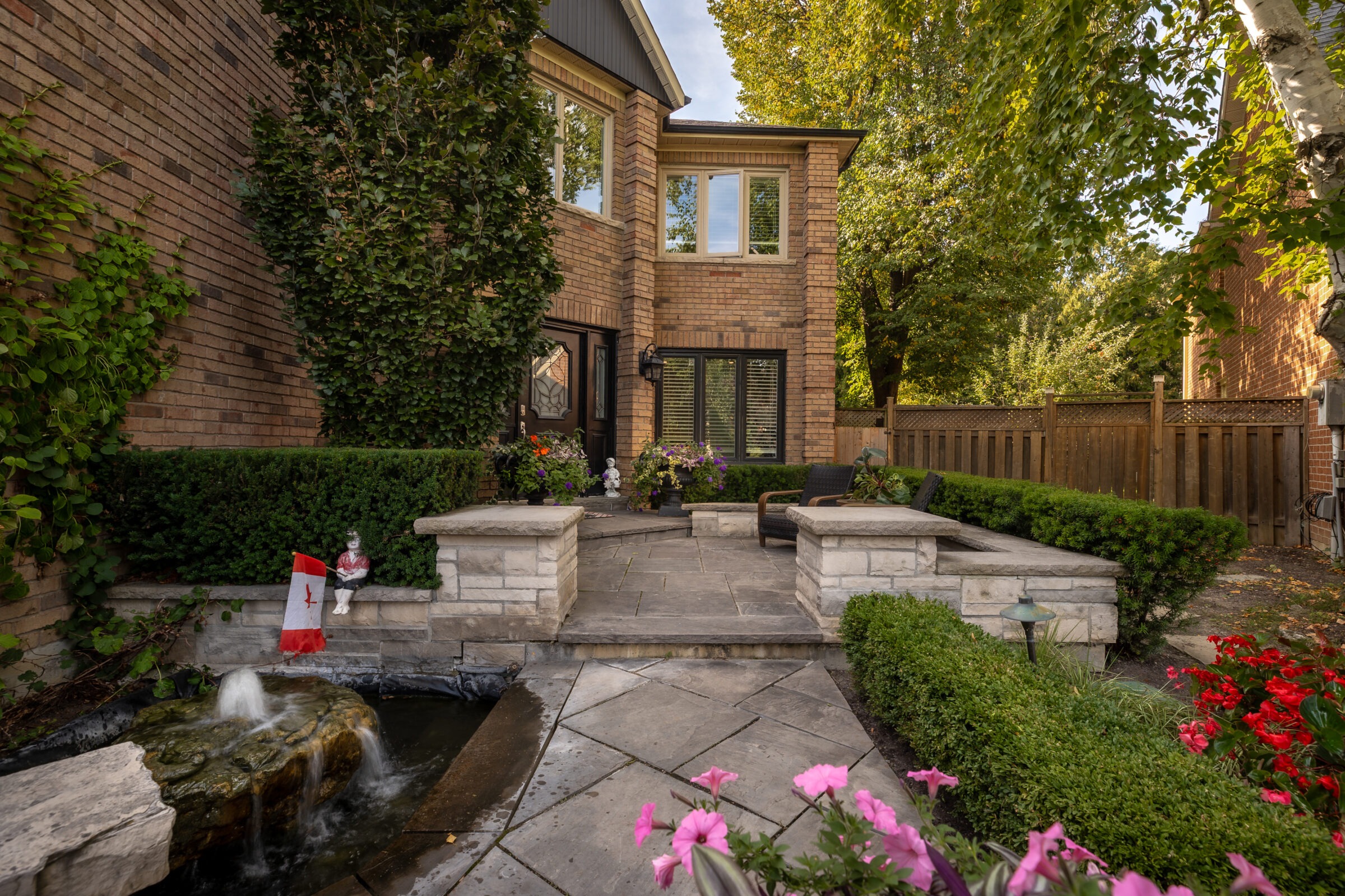 Cozy brick house with landscaped garden, small waterfall, and Canadian flag. Surrounded by lush trees, flowers, and elegant stone walkway.