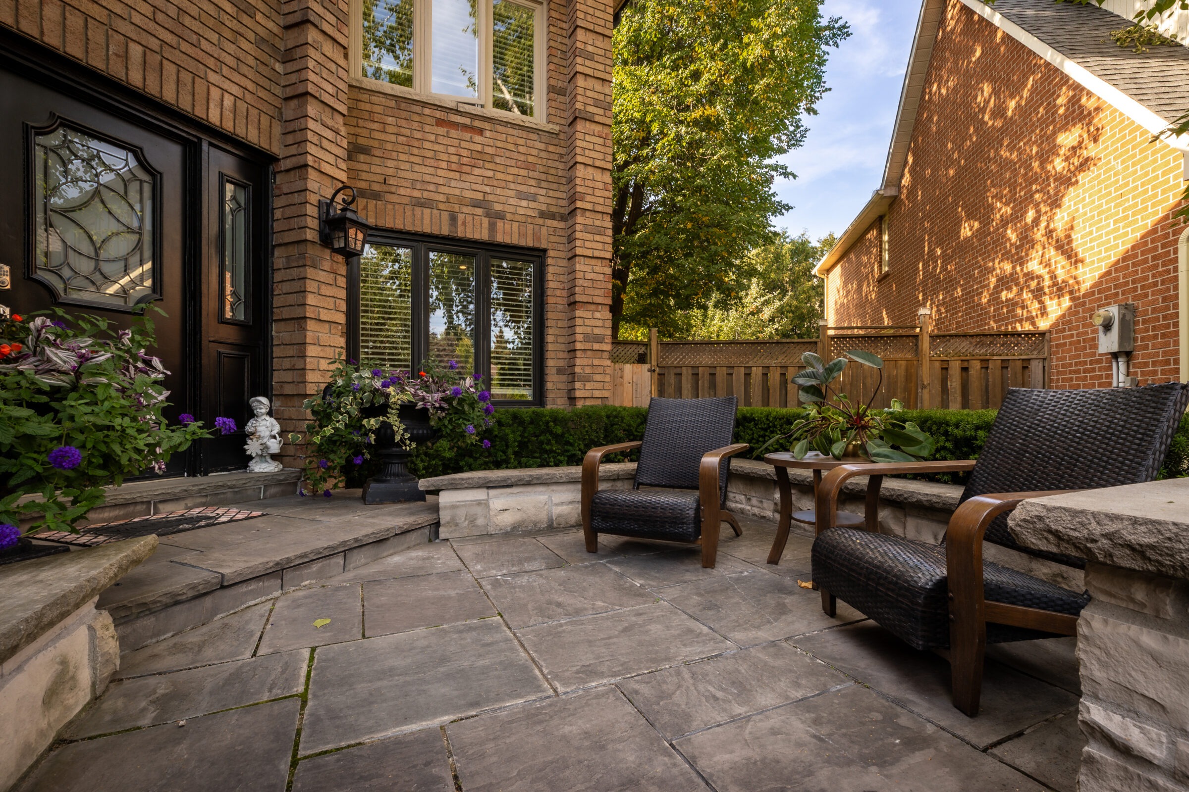 Brick house with a cozy patio features two wicker chairs, potted plants, and trees, creating a peaceful and inviting outdoor space.