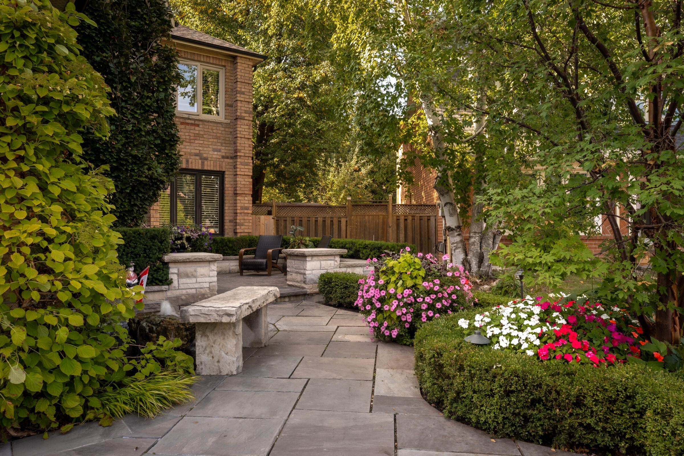 A serene garden with stone paths, lush greenery, colorful flowers, and a brick house in the background, offering a peaceful seating area.