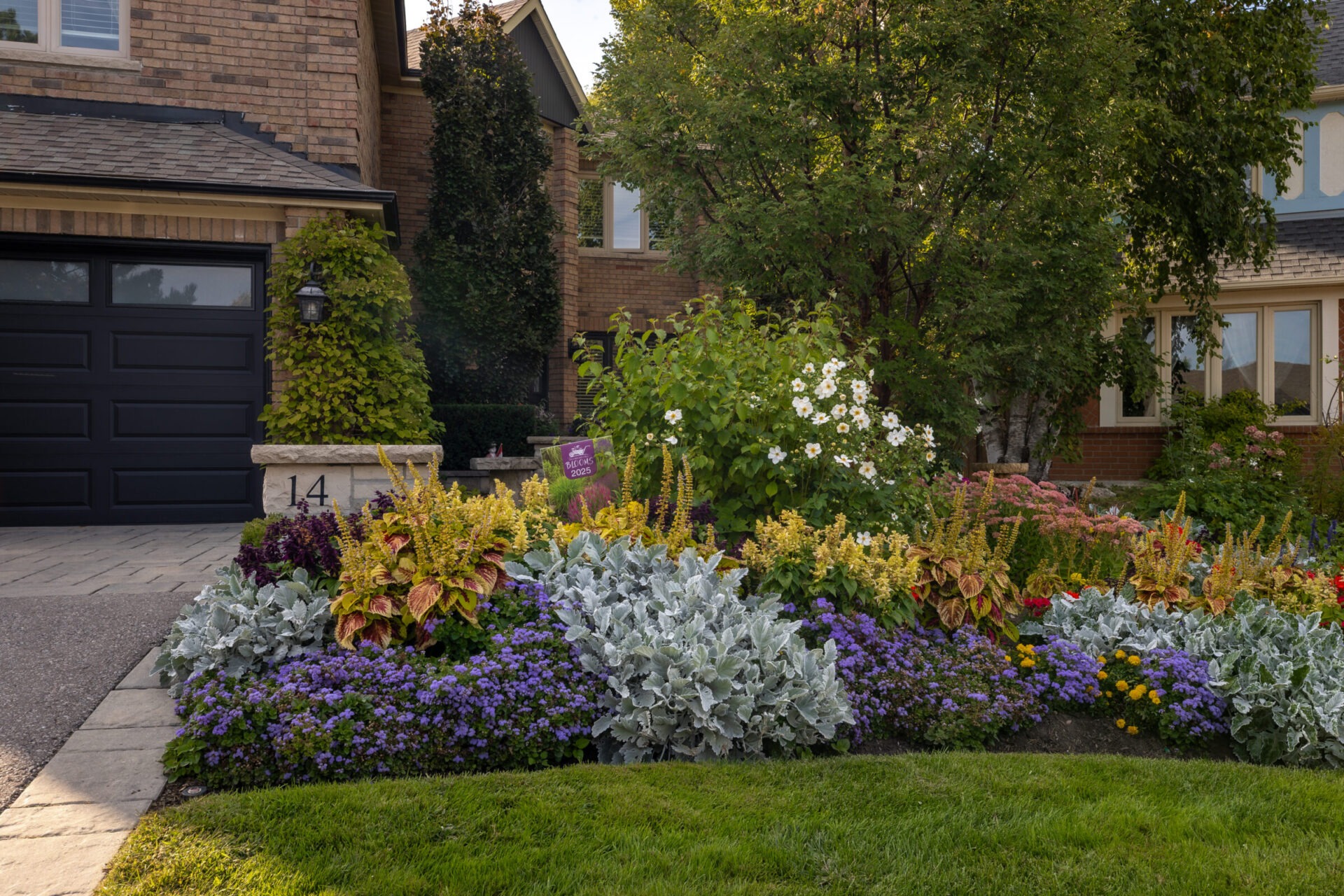 A well-maintained garden with colorful flowers in front of a brick house, featuring a garage and driveway. No recognizable landmarks present.