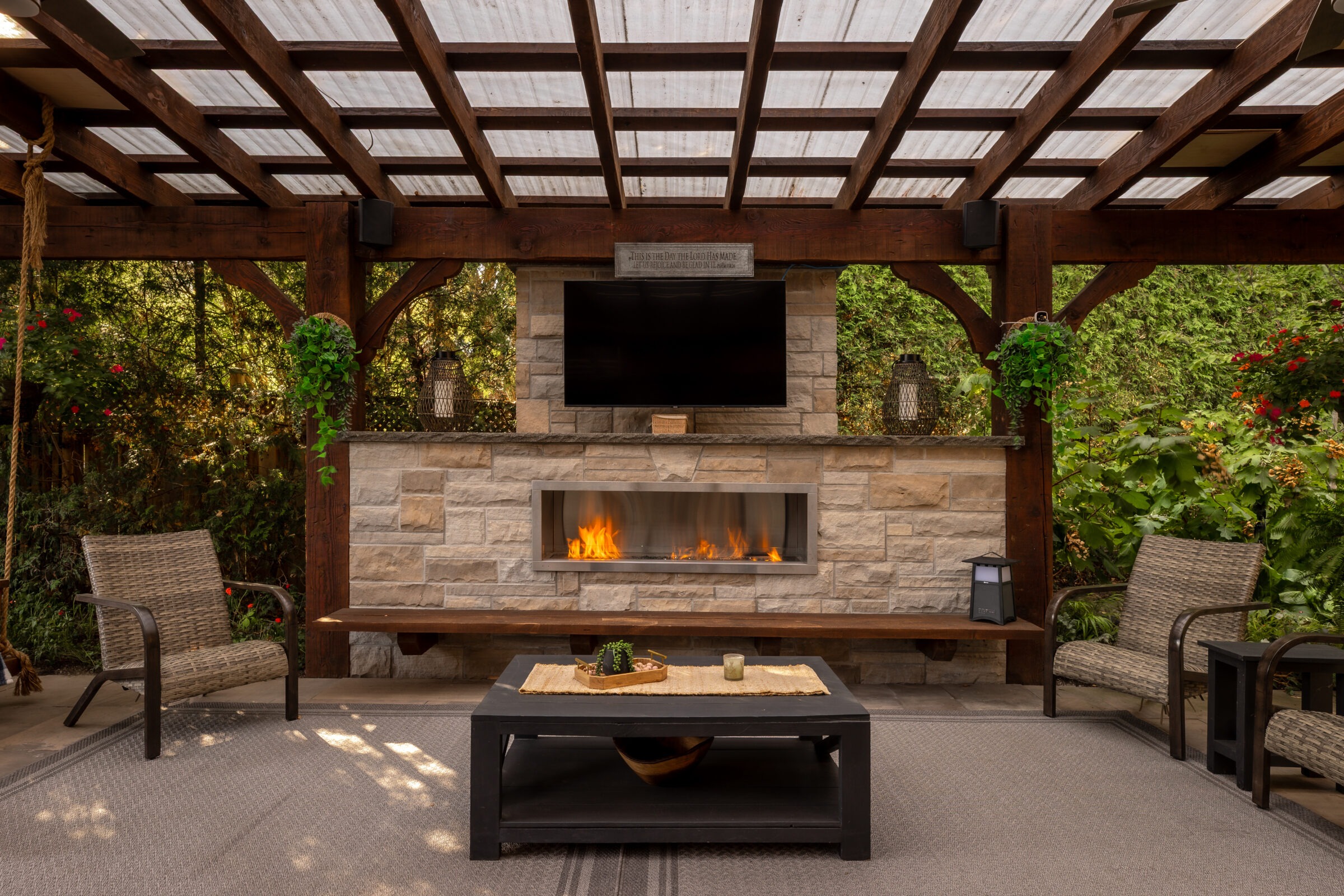Cozy patio with wicker chairs, modern fireplace, and mounted TV under a wooden pergola. Lush greenery surrounds the inviting outdoor space.