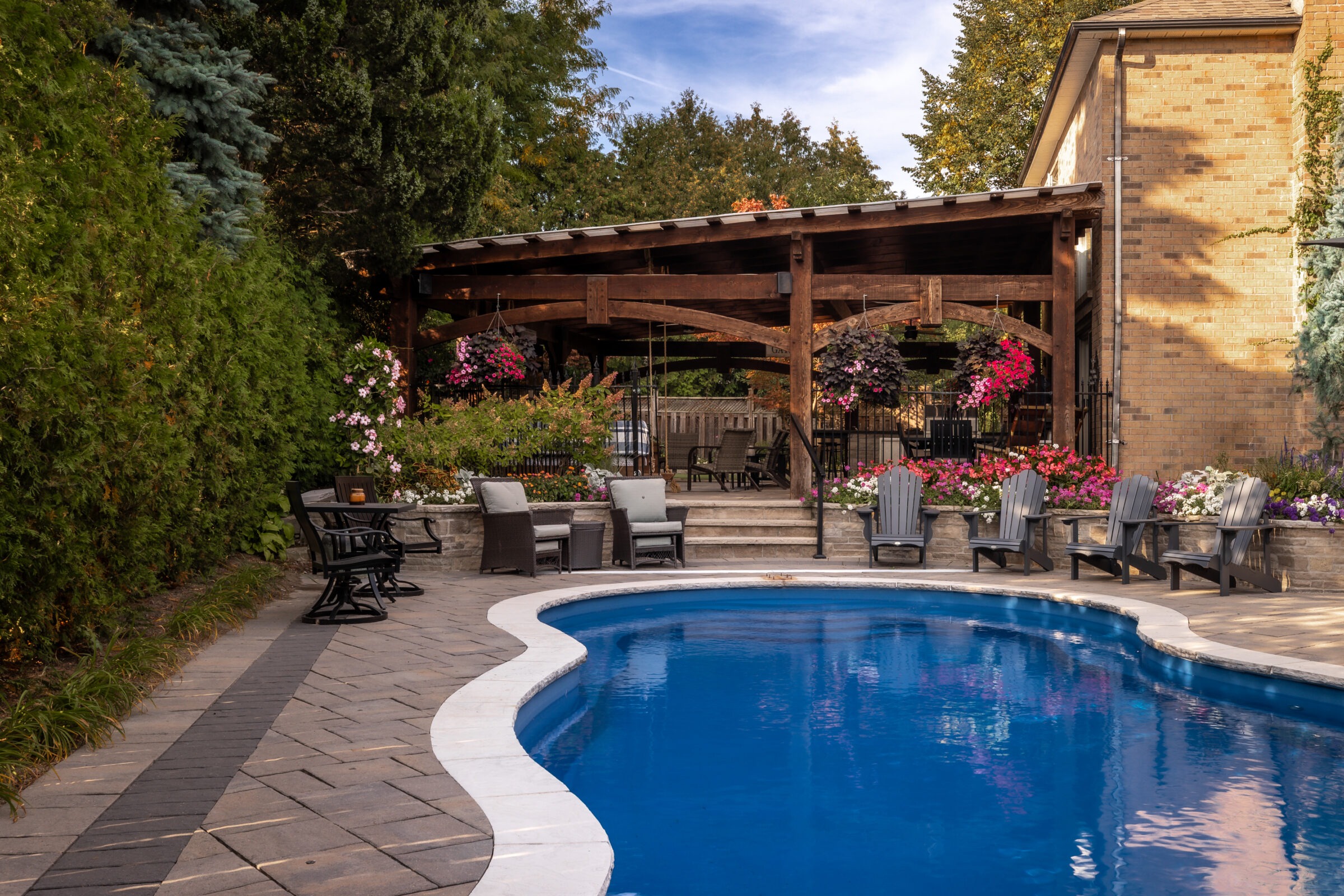 A tranquil backyard with a curved pool, wooden pergola, and patio chairs surrounded by lush greenery and colorful flowers, under a clear blue sky.