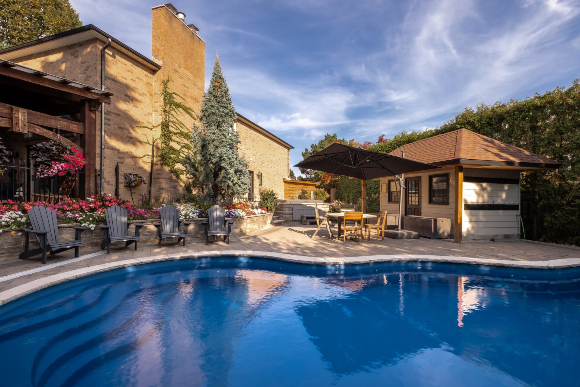 A tranquil backyard features a curved pool, patio chairs, parasol, and a small building, surrounded by lush greenery and brick facades.