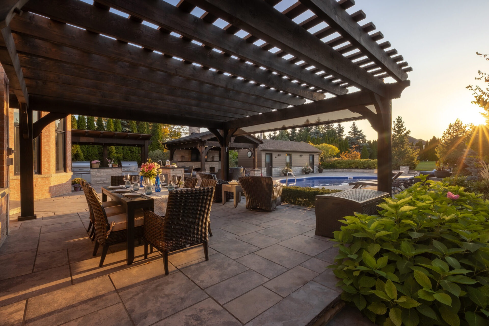 A stylish outdoor dining area under a pergola, adjacent to a pool with landscaped garden. Sun setting, casting warm light across the patio.