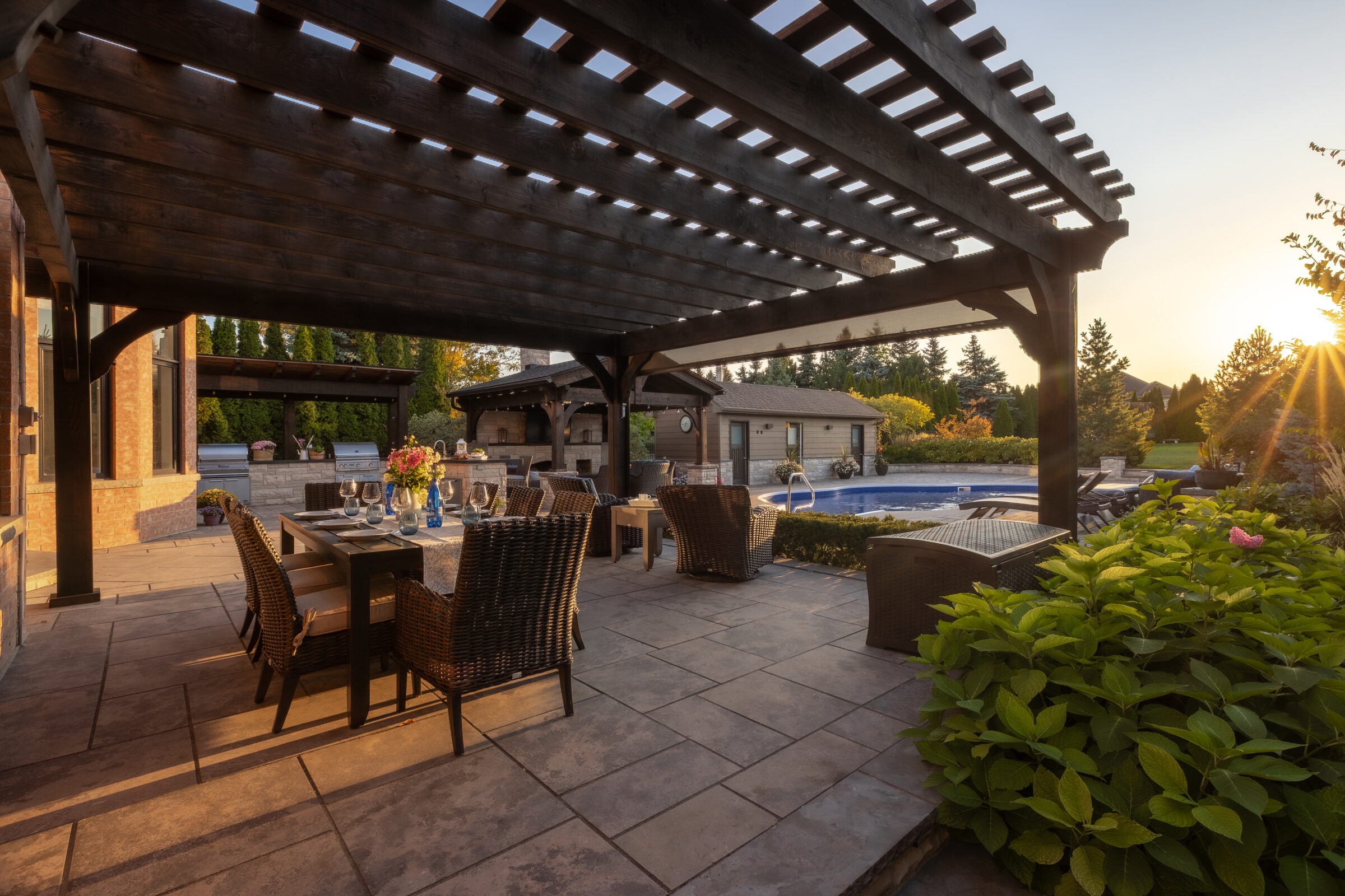 A stylish outdoor dining area under a pergola, adjacent to a pool with landscaped garden. Sun setting, casting warm light across the patio.