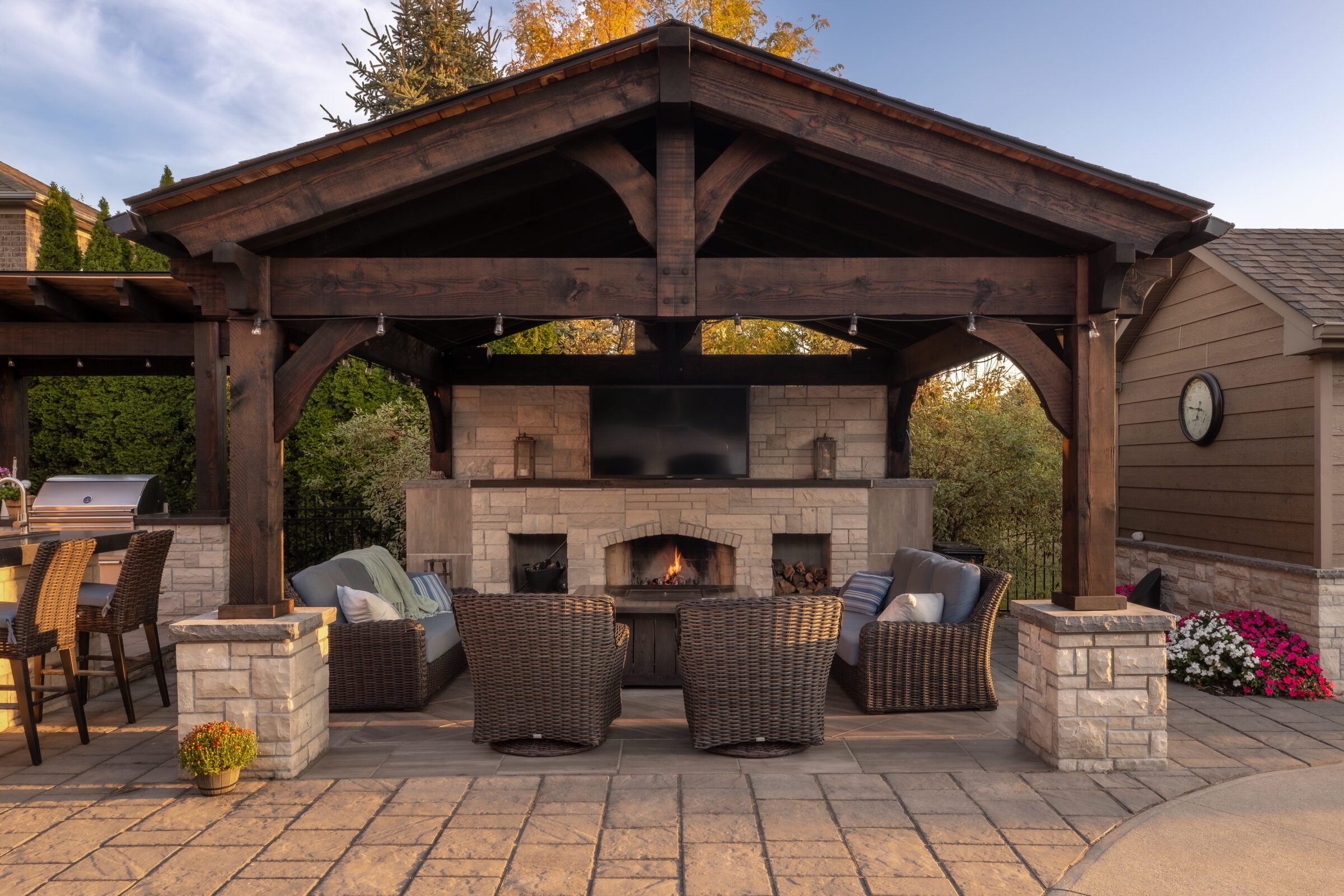 A cozy outdoor seating area with wicker chairs and a stone fireplace beneath a wooden pergola, surrounded by lush greenery and warm lighting.