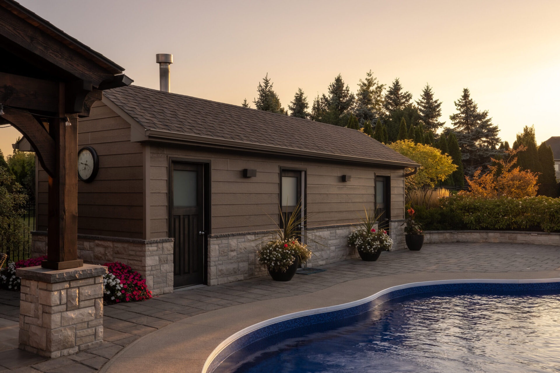 A peaceful backyard features a pool, wooden pool house, and potted plants, surrounded by lush trees under a warm evening sky.