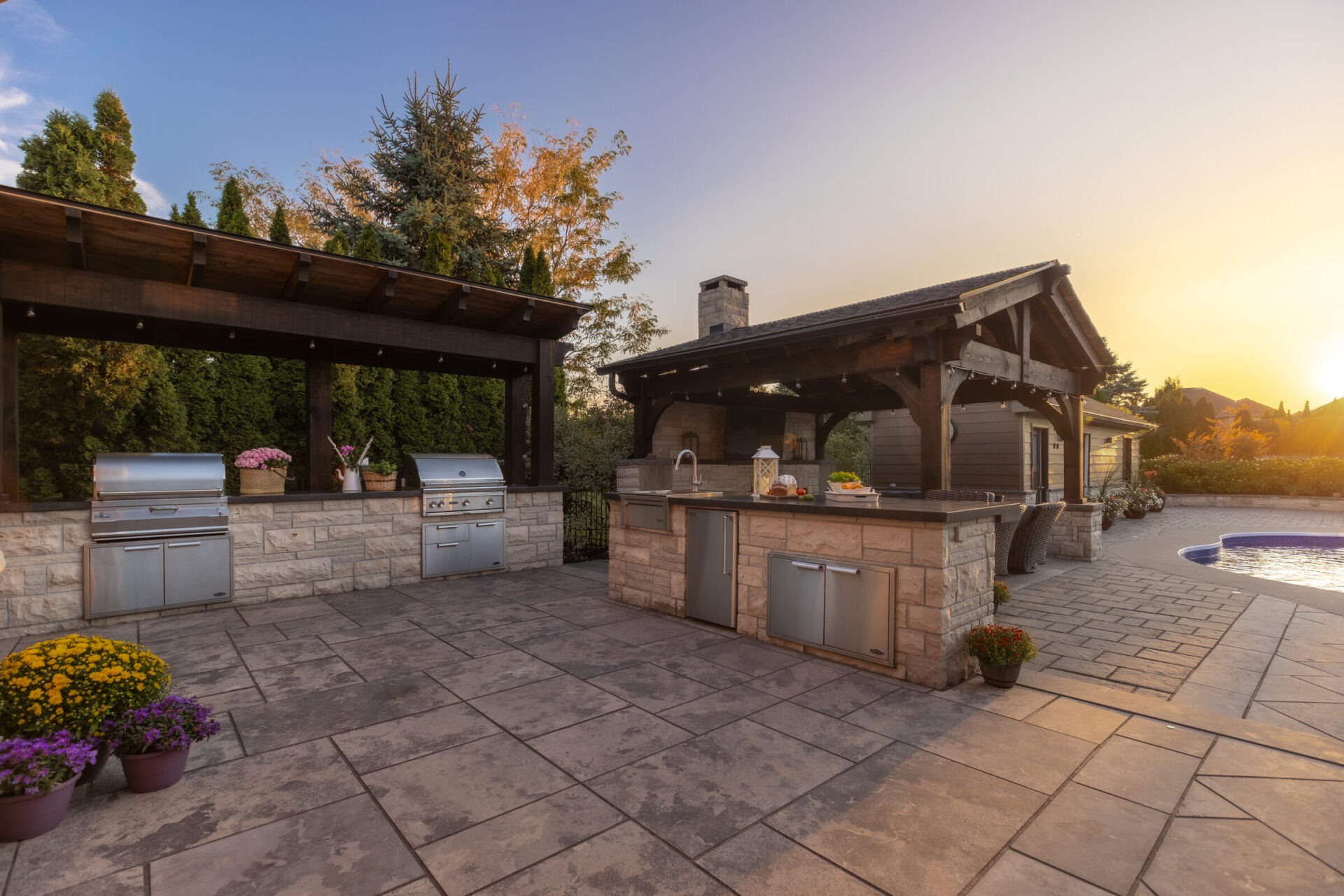 Outdoor kitchen with stone countertops, stainless steel appliances, and a pergola. Flower pots decorate the spacious patio beside a pool at sunset.