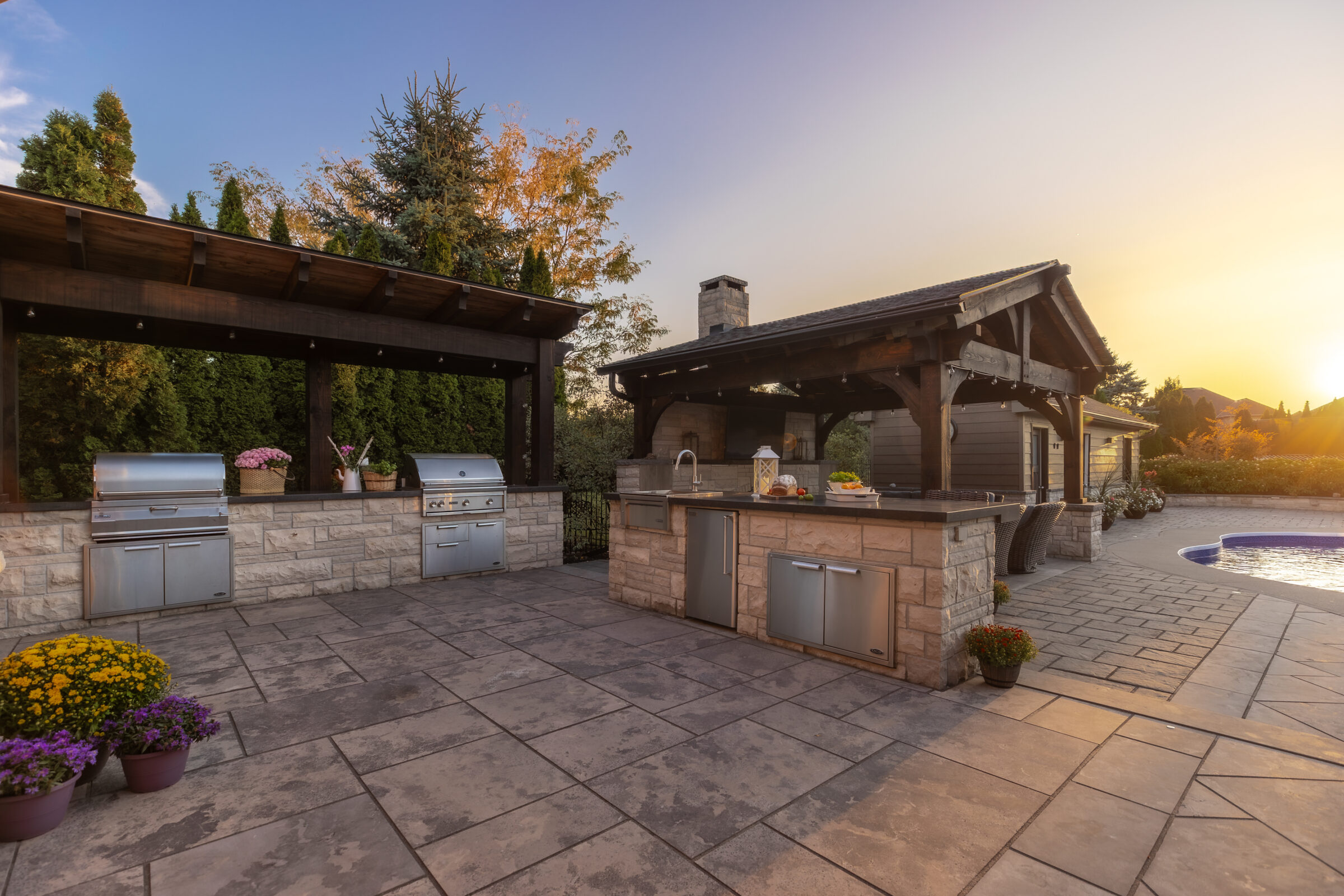 Outdoor kitchen with stone countertops, stainless steel appliances, and a pergola. Flower pots decorate the spacious patio beside a pool at sunset.