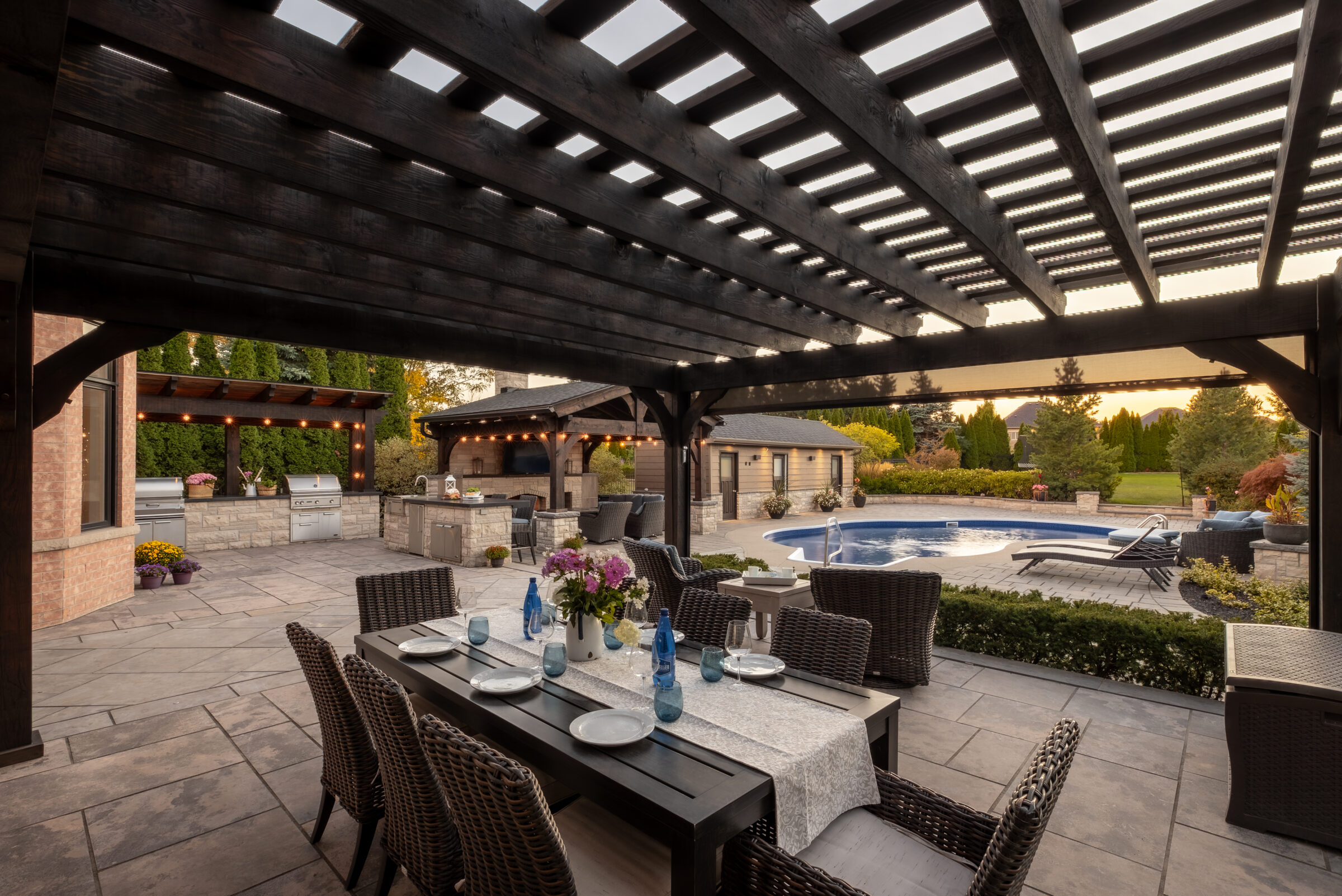 Patio features pergola, dining table with place settings, poolside view, and lush garden. Outdoor kitchen in the background enhances inviting atmosphere.