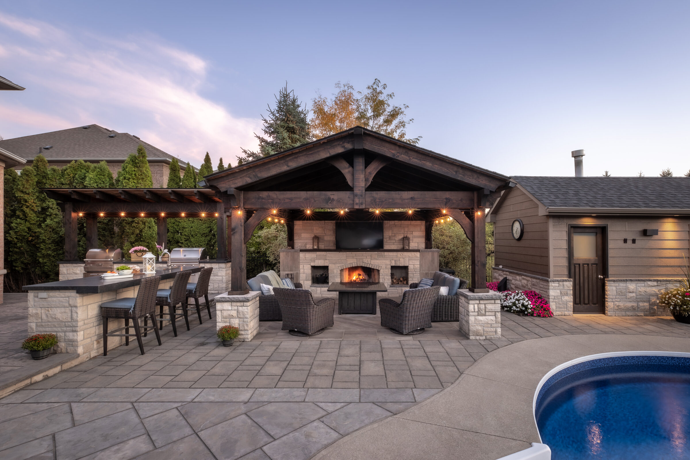 A backyard features a pool, covered patio with a TV, fireplace, seating area, bar counter, and ambient lighting against a twilight sky.