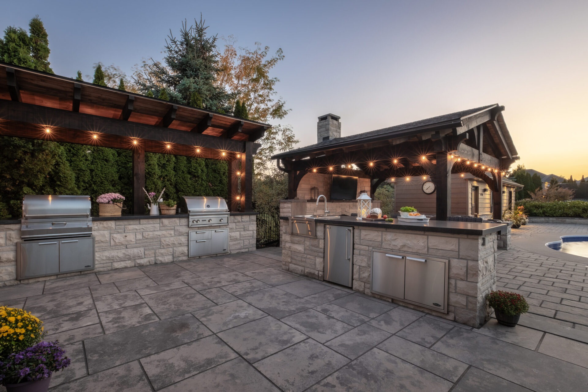 Outdoor kitchen area at sunset, featuring two grilling stations, stone countertops, string lights, and surrounding trees on a paved patio.