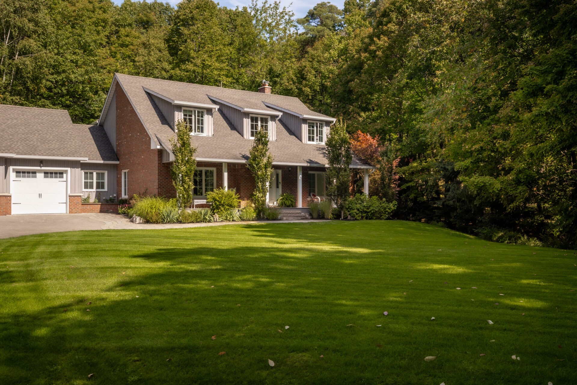 Brick house with gable roof, surrounded by lush trees. Green lawn extends in front, creating a serene, secluded residential setting.