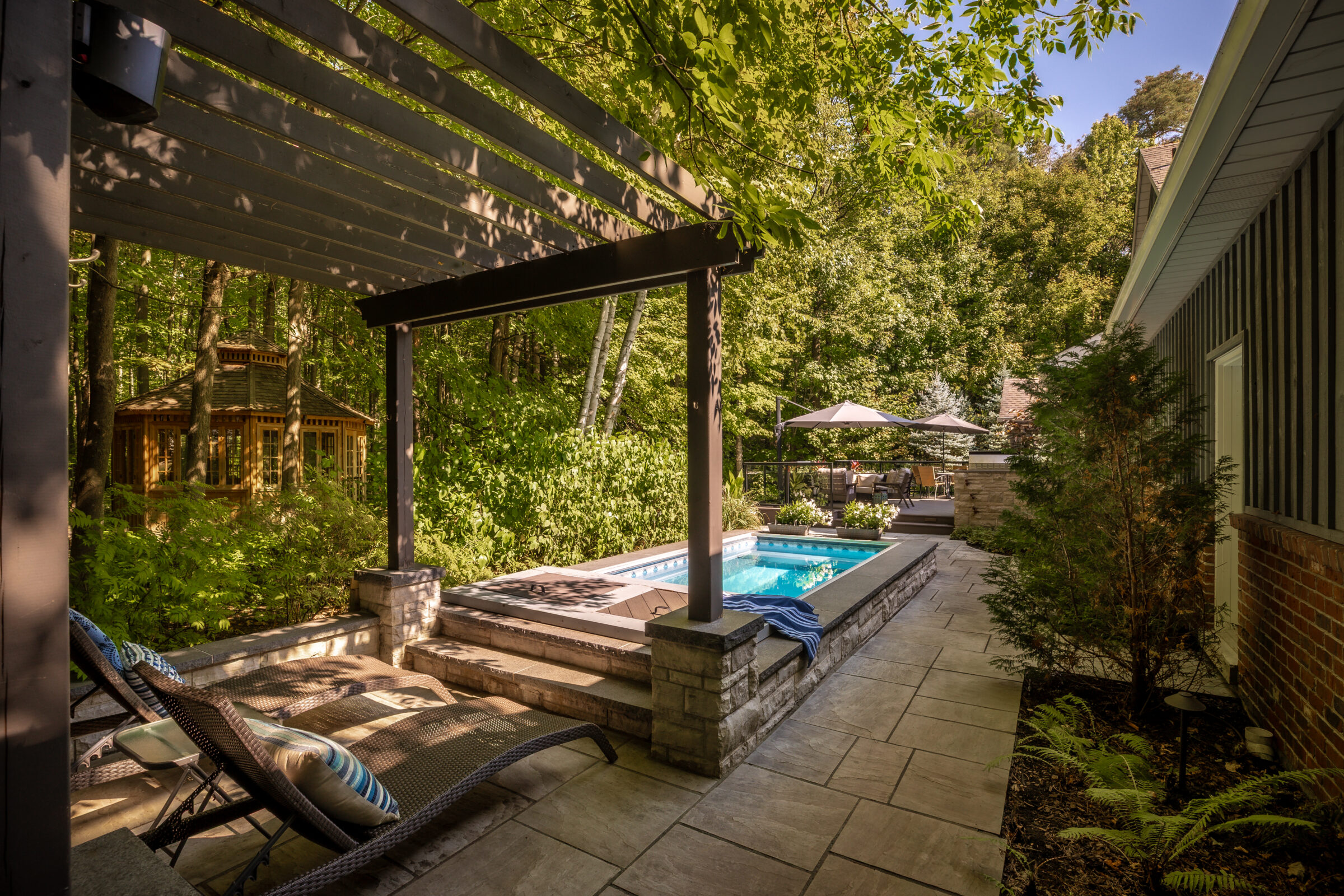 A tranquil backyard scene features a small pool, lounge chairs, pergola, gazebo, and lush greenery surrounding the area, creating a peaceful retreat.