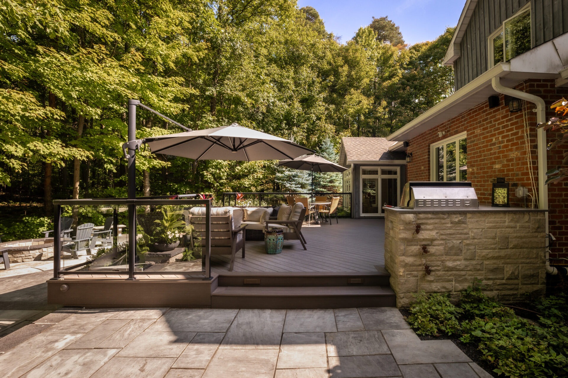 A spacious backyard patio with outdoor seating, a large umbrella, and a grill, surrounded by lush trees and a brick house.