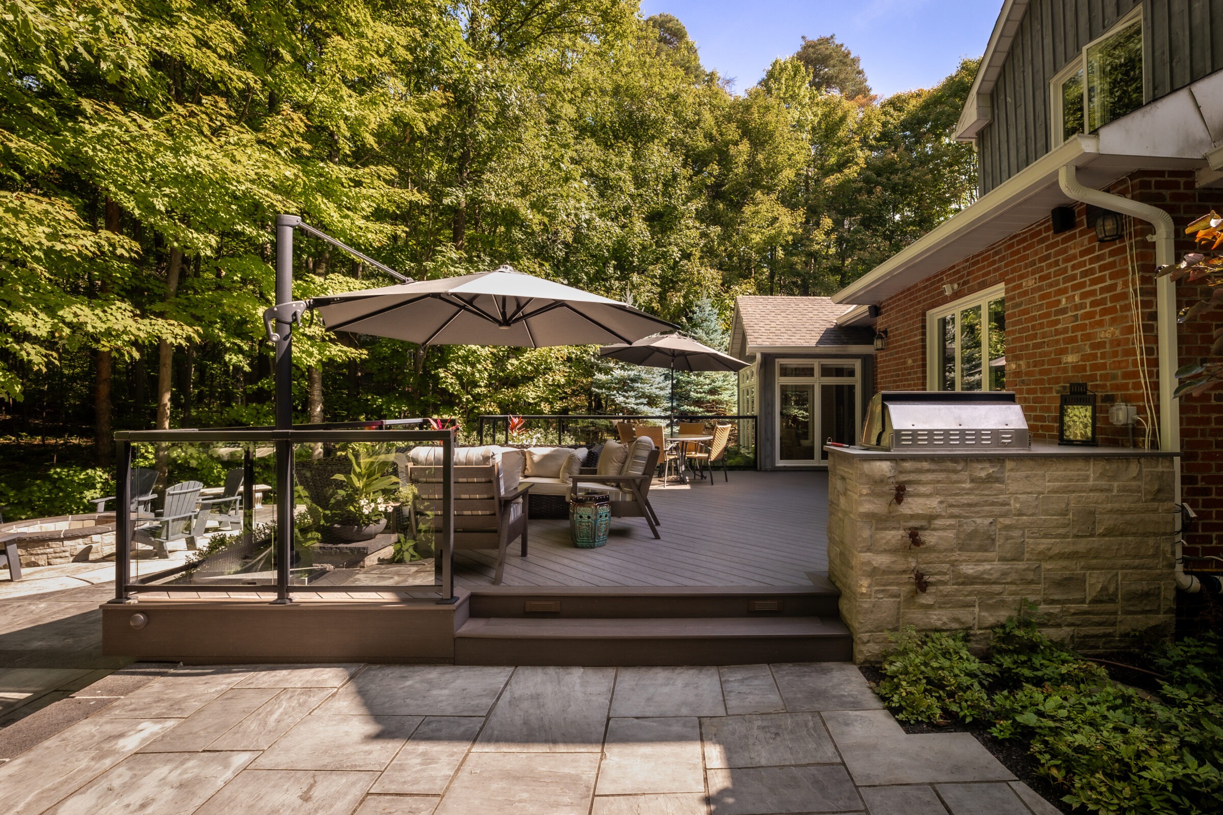 A spacious backyard patio with outdoor seating, a large umbrella, and a grill, surrounded by lush trees and a brick house.