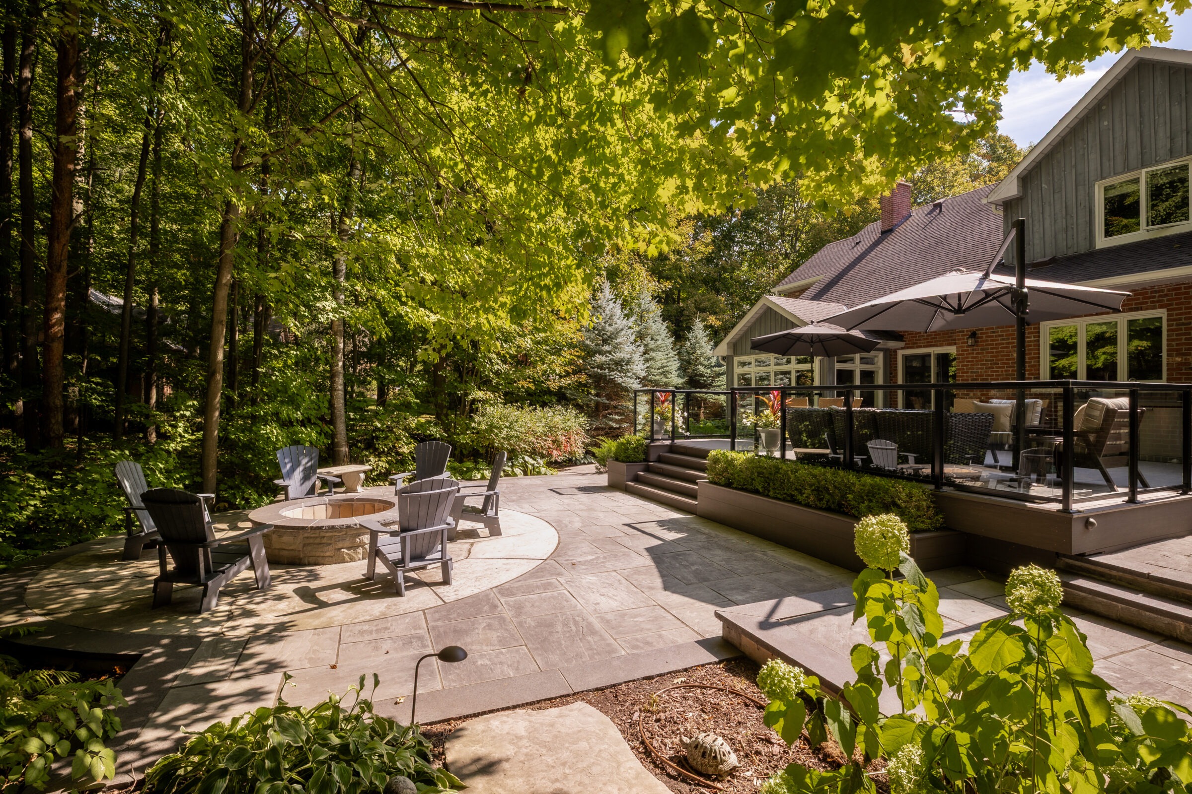 A peaceful backyard with patio furniture, a fire pit, surrounded by lush trees and garden, attached to a modern house with large windows.