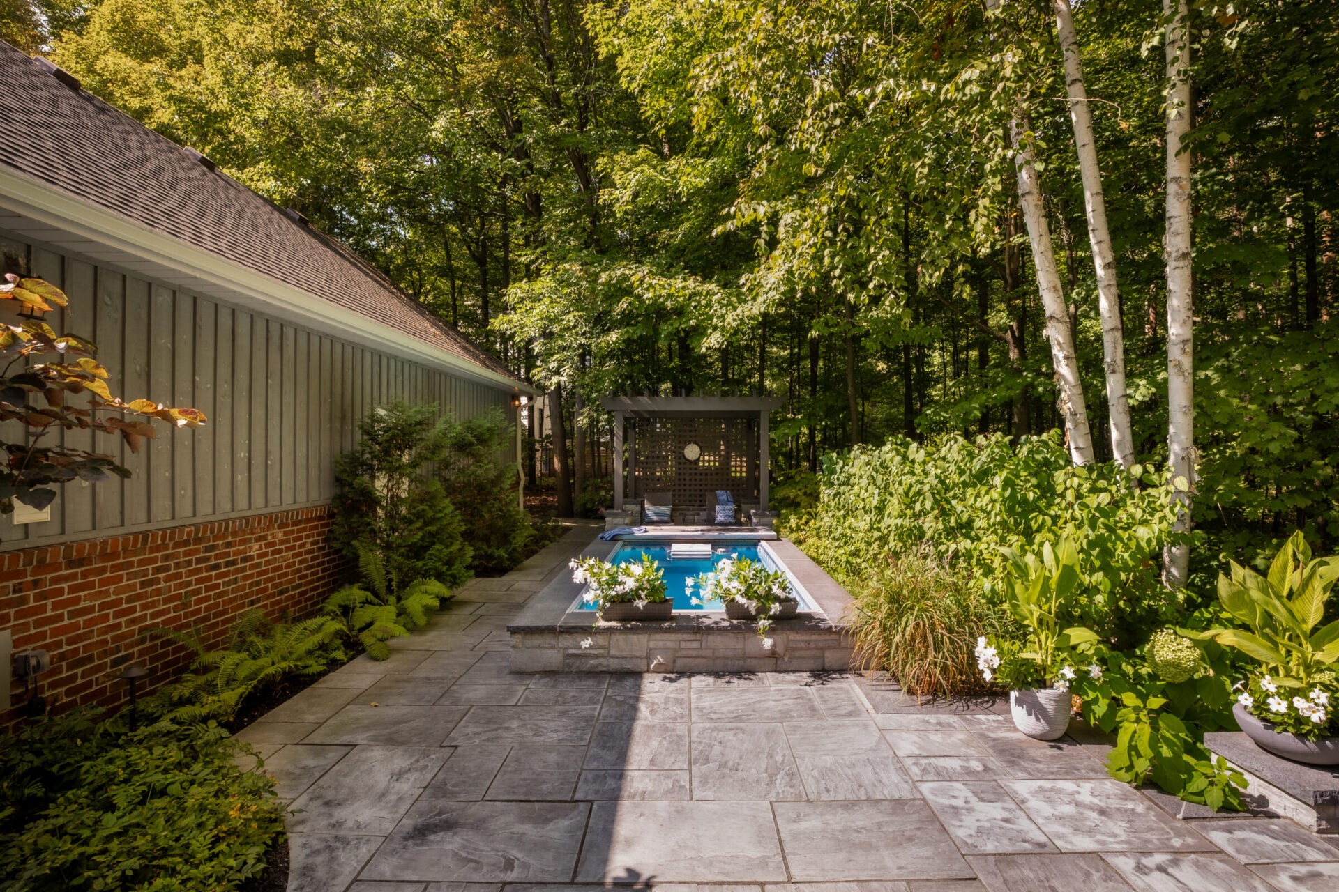 A serene backyard features a small pool, stone patio, lush greenery, and wooden pergola, surrounded by trees and adjacent to a brick house.