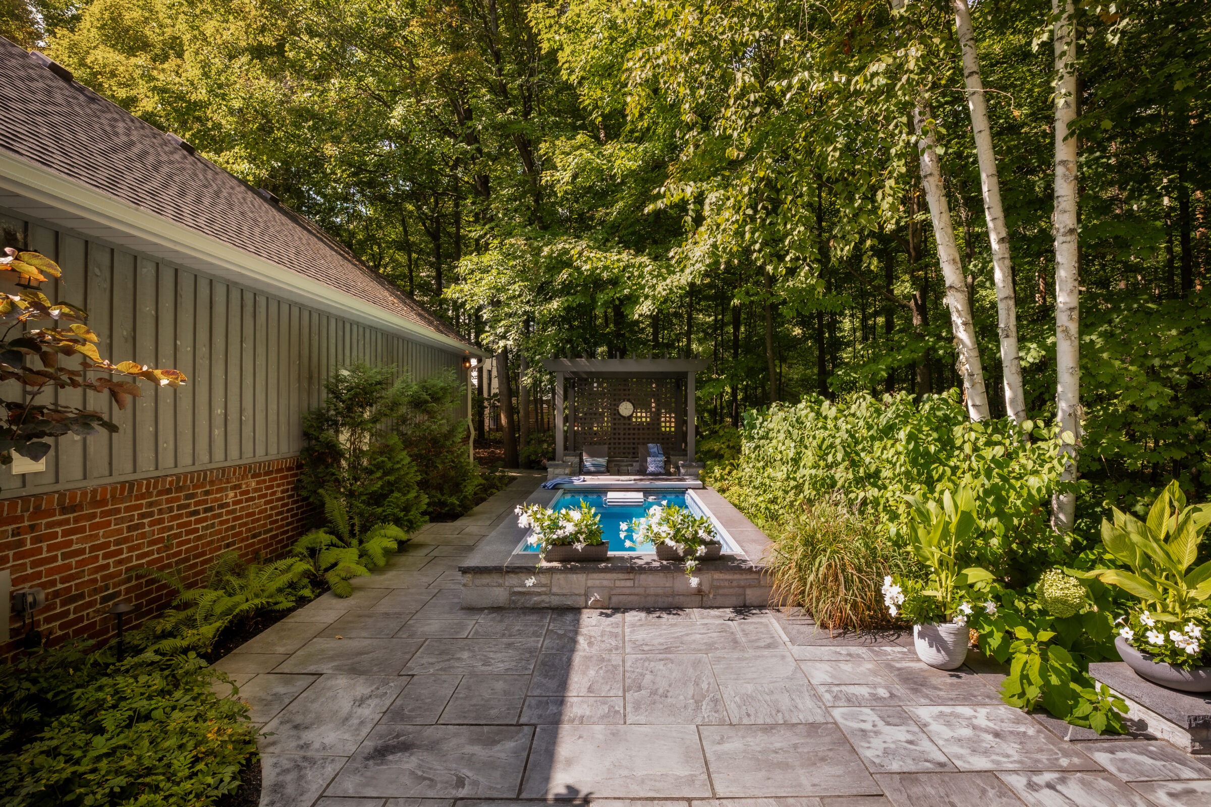 A serene backyard features a small pool, stone patio, lush greenery, and wooden pergola, surrounded by trees and adjacent to a brick house.