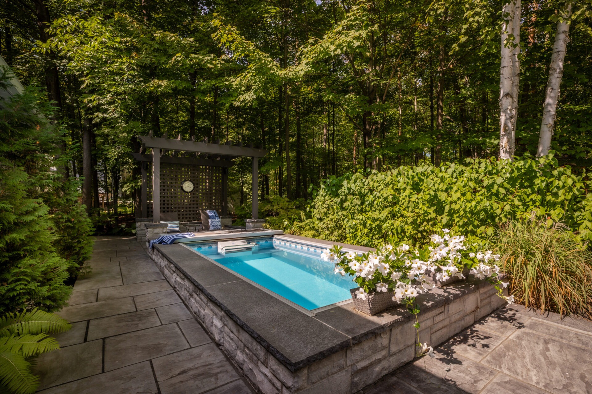 A serene backyard setting with a small pool, surrounded by lush greenery. A pergola with chairs provides a relaxing spot nearby.